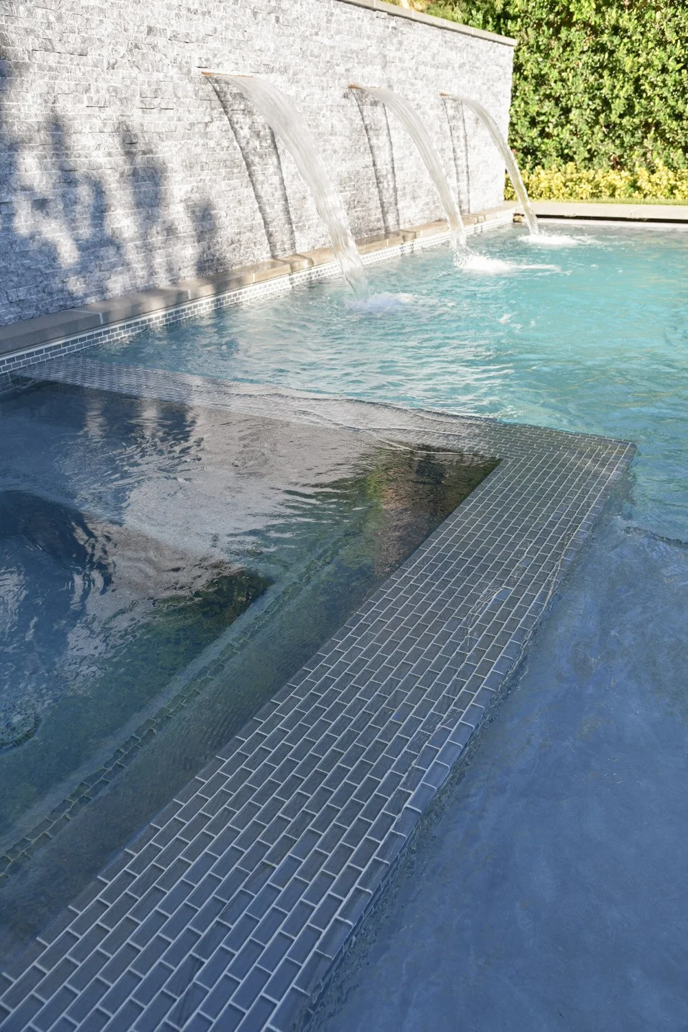 Close view of a tiled spa divider and calm water beside a rectangular pool with a stone waterfall wall.