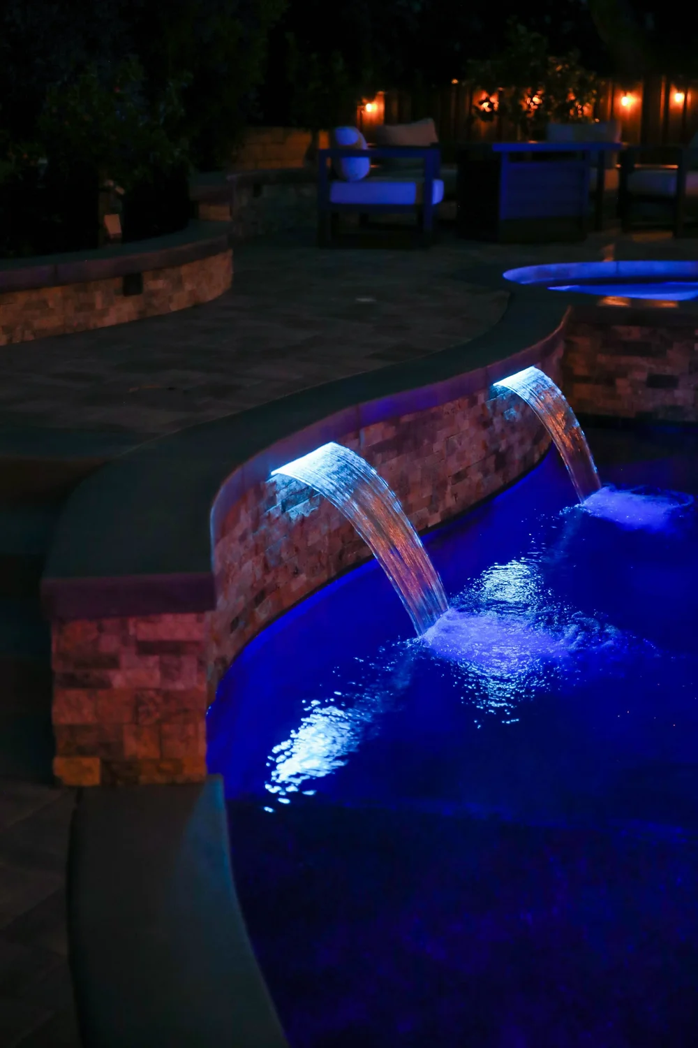 Two lit spillways pour from a curved stone wall into a blue-lit pool at night.