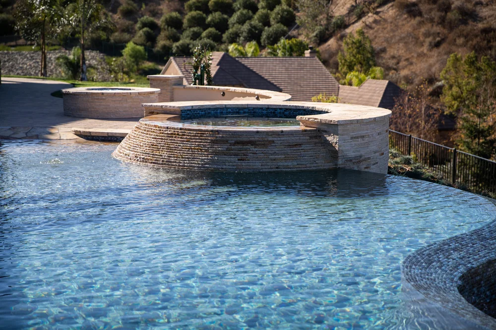 Stone-faced circular spa above a blue-tiled pool, with a planted hillside beyond the water.