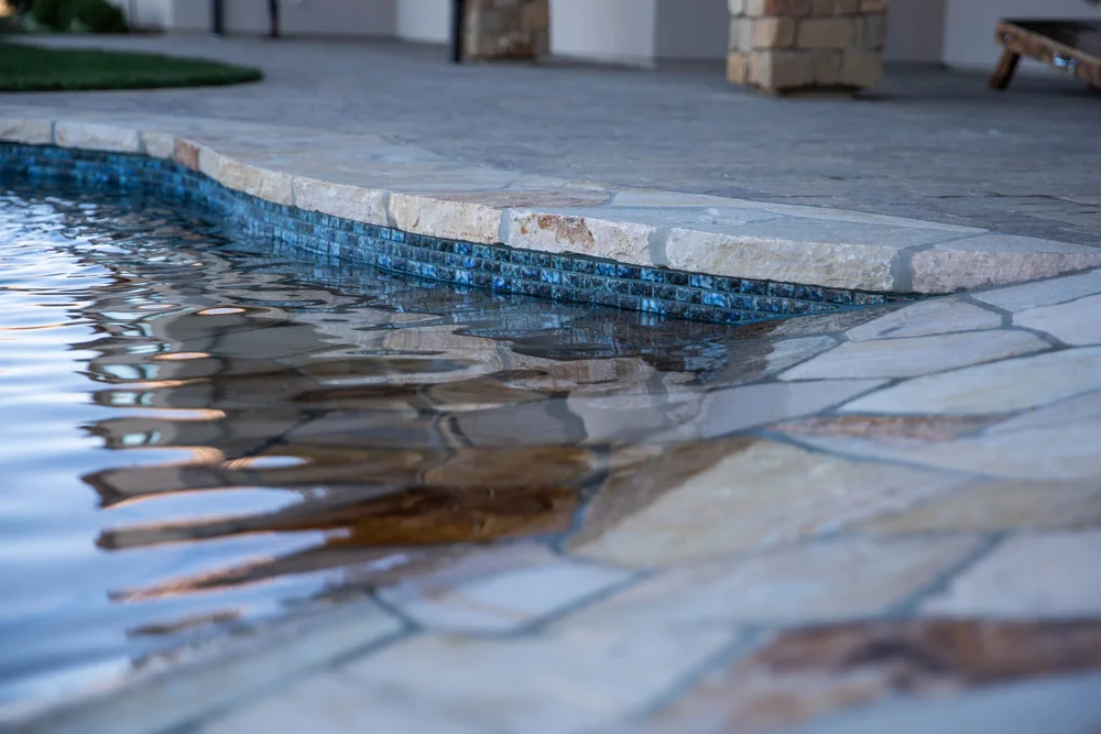 Close view of a shallow beach entry with stone paving and blue mosaic tile along the curved pool wall.