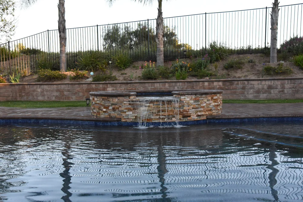 Water flowing from a raised round stone-faced spa into a curved pool below a planted retaining wall.