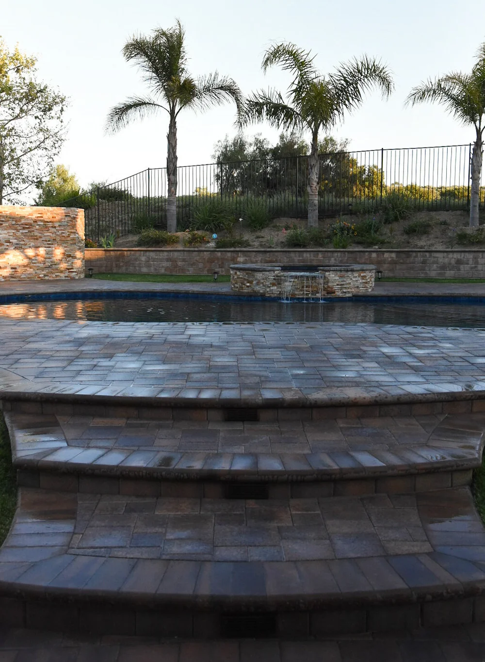 Broad curved paver steps leading toward a pool, raised stone-faced spa and planted slope with palms.