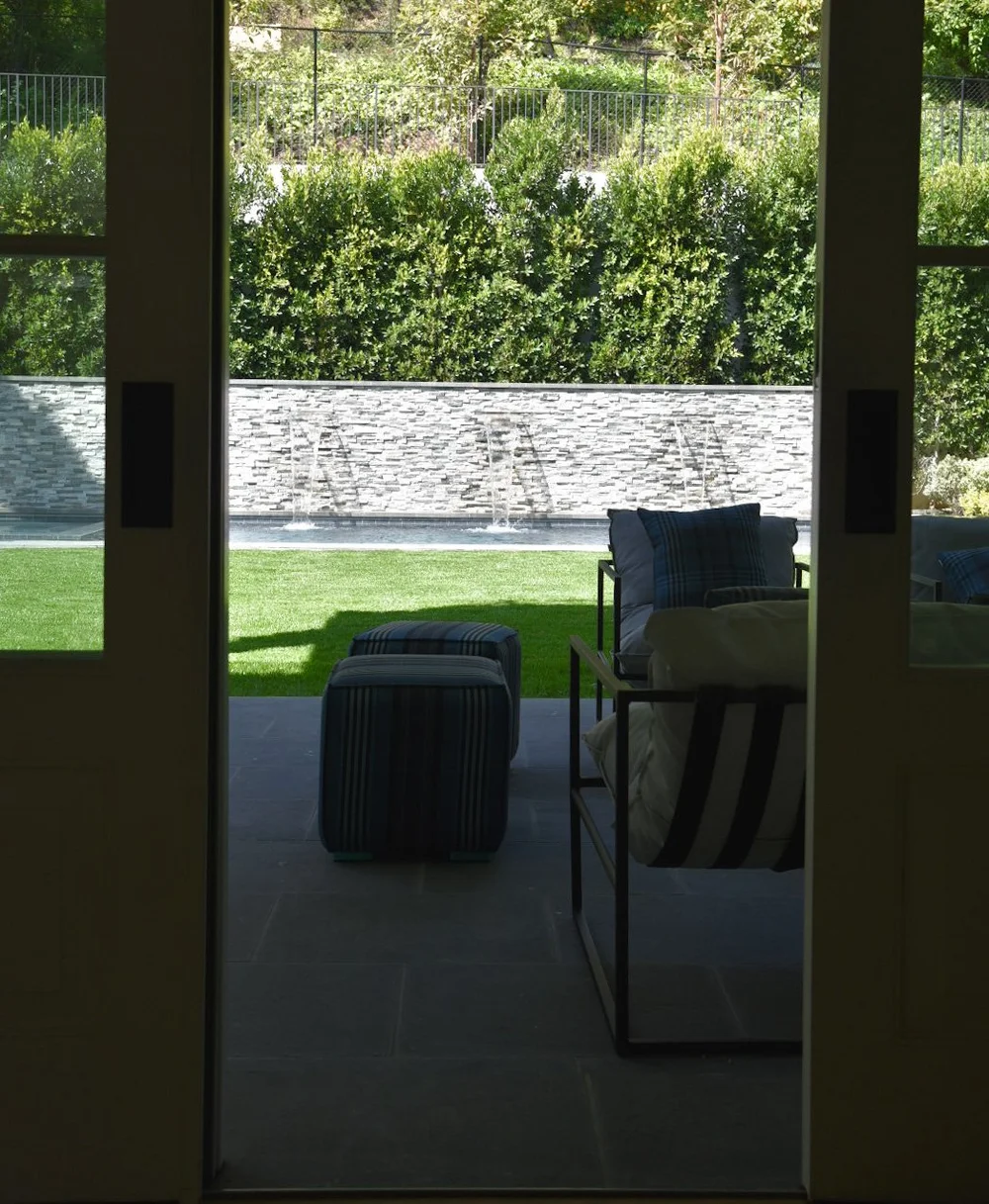 View through open patio doors toward outdoor seating, a lawn and a pool with a long stone water wall.