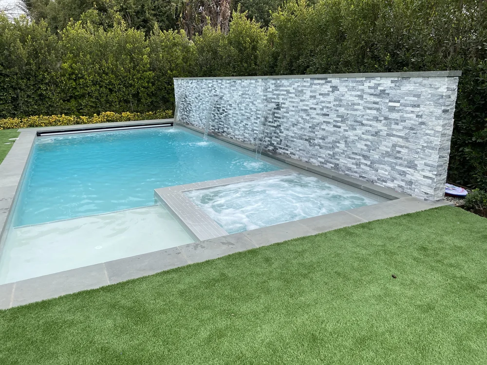 A rectangular turquoise pool includes a bubbling square spa and shallow entry area beside a tall textured wall.