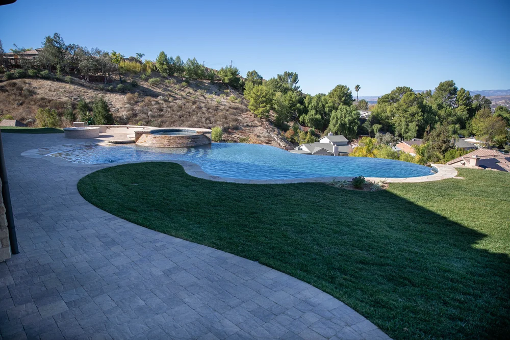 Wide view of a curved infinity-edge pool and raised spa beside a lawn and paved patio.
