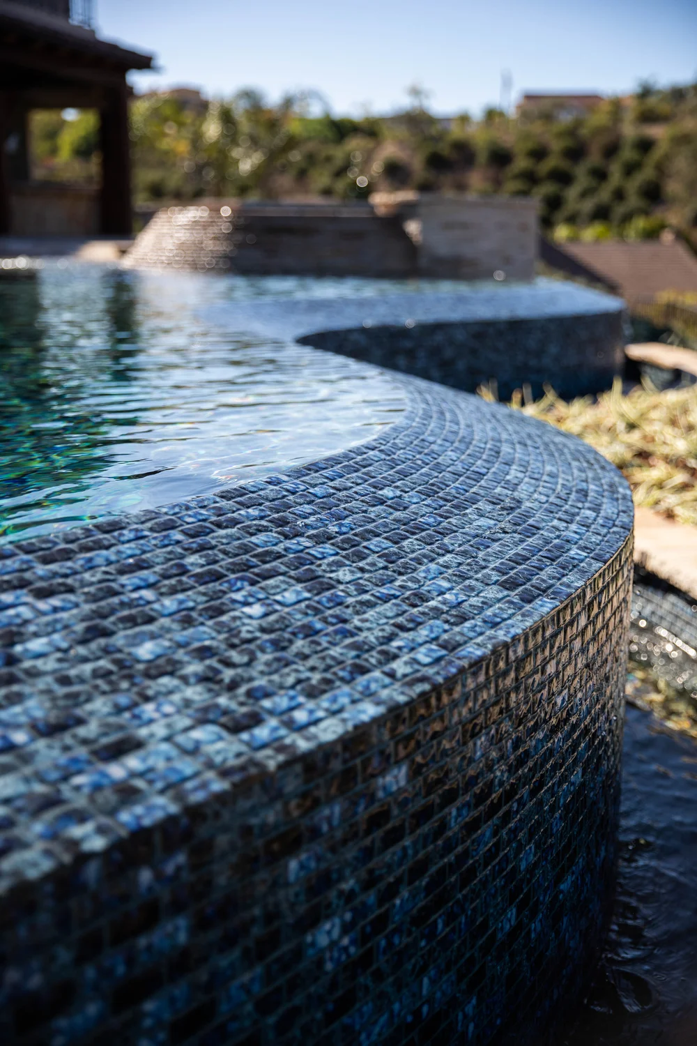 Close-up of small blue mosaic tiles along a curved infinity edge, with the spa softly blurred in the background.