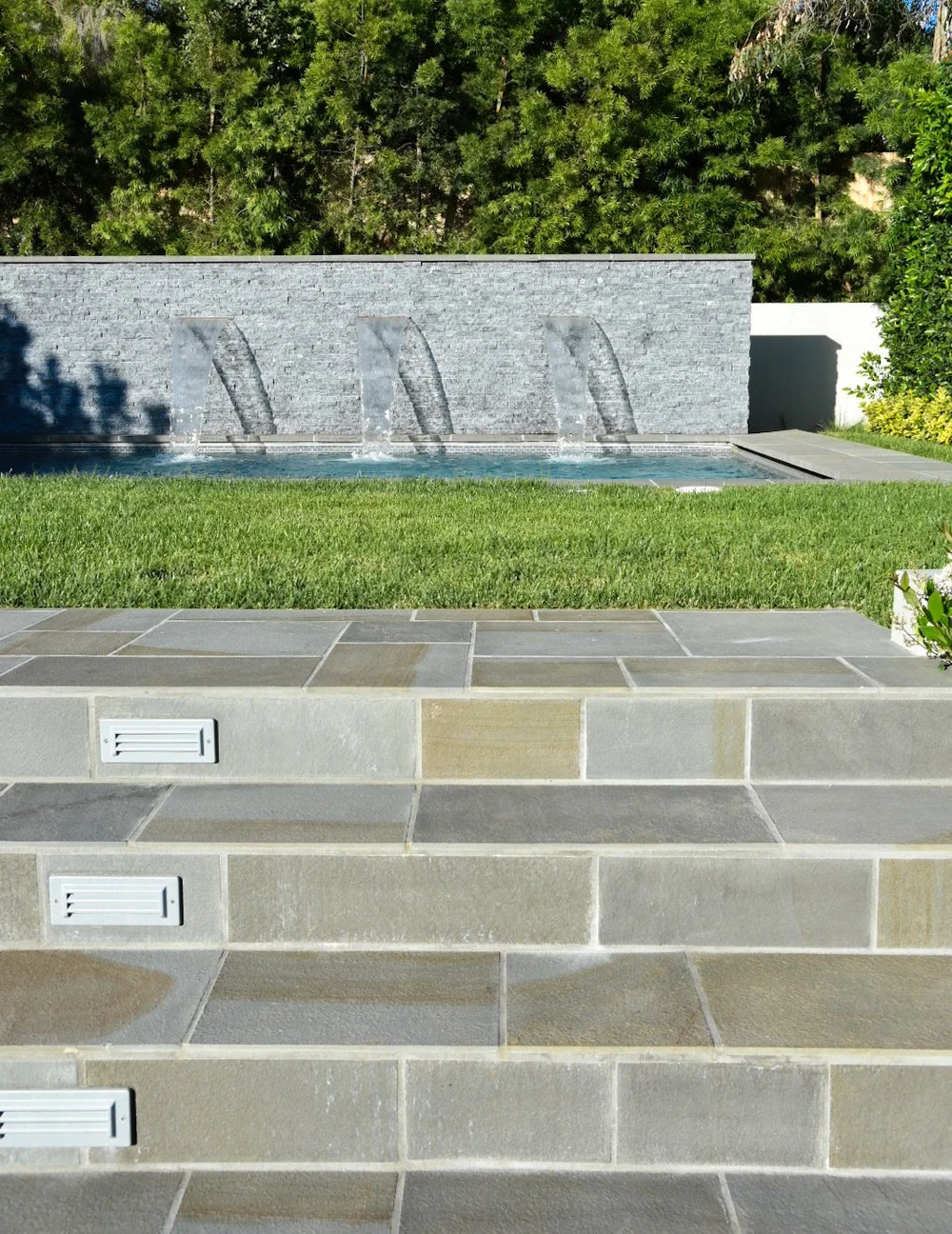 Tiled outdoor steps with inset step lights leading past a lawn toward a pool and stone water wall.