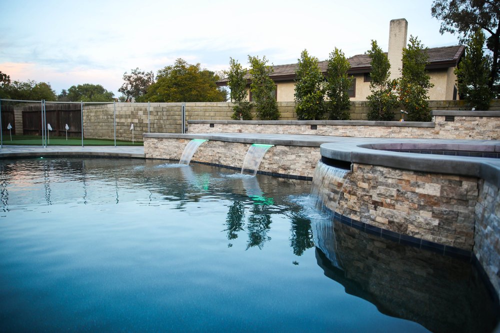 Low view across a pool toward a raised round stone-faced spa and two spillways in the adjoining wall.