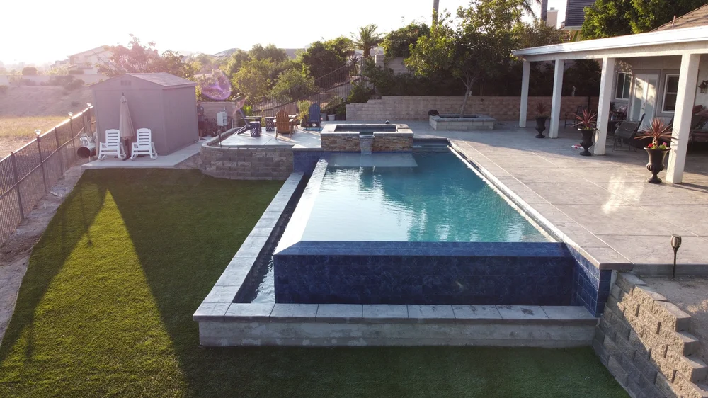 A long rectangular pool runs beside a covered patio, with a blue-tiled overflow edge, lawn, and raised spa.