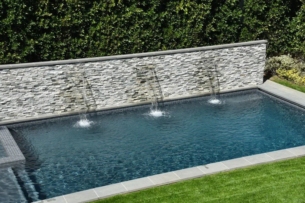 Elevated view of a rectangular pool with three spillways in a stacked-stone wall, bordered by lawn and light coping.