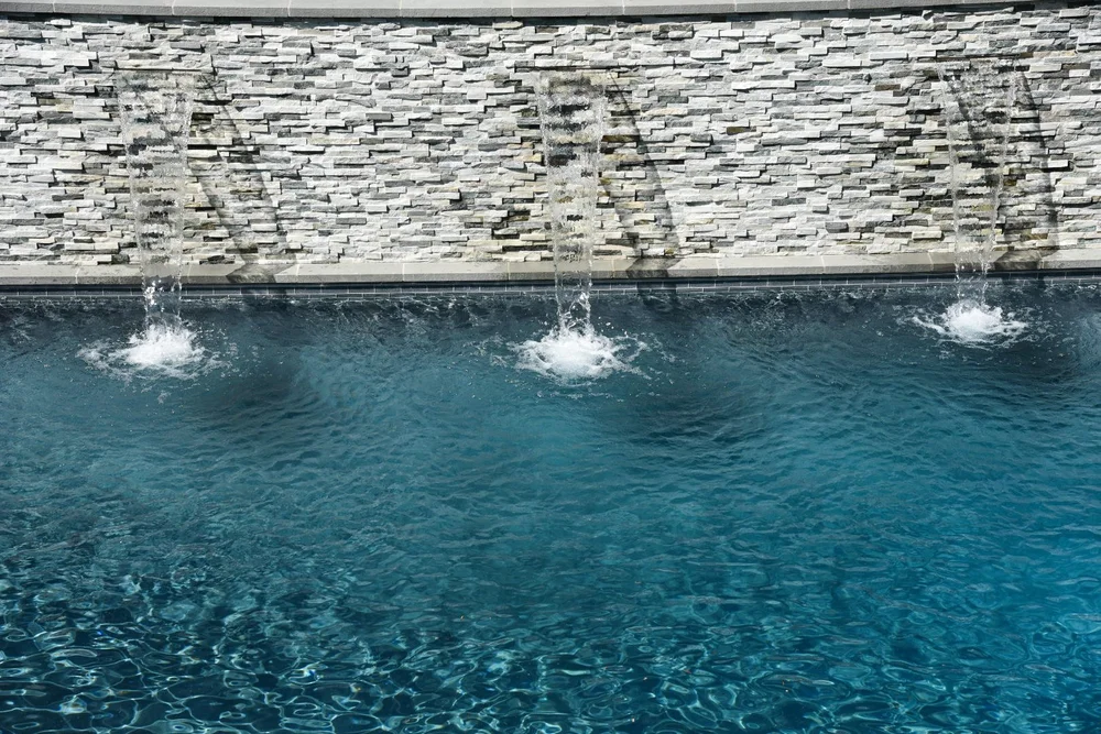 Three streams of water falling from a long stacked-stone feature wall into blue pool water.