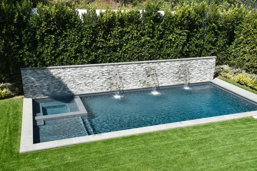 Rectangular pool and square integrated spa beside a lawn, with three wall spillways and a tall hedge behind.
