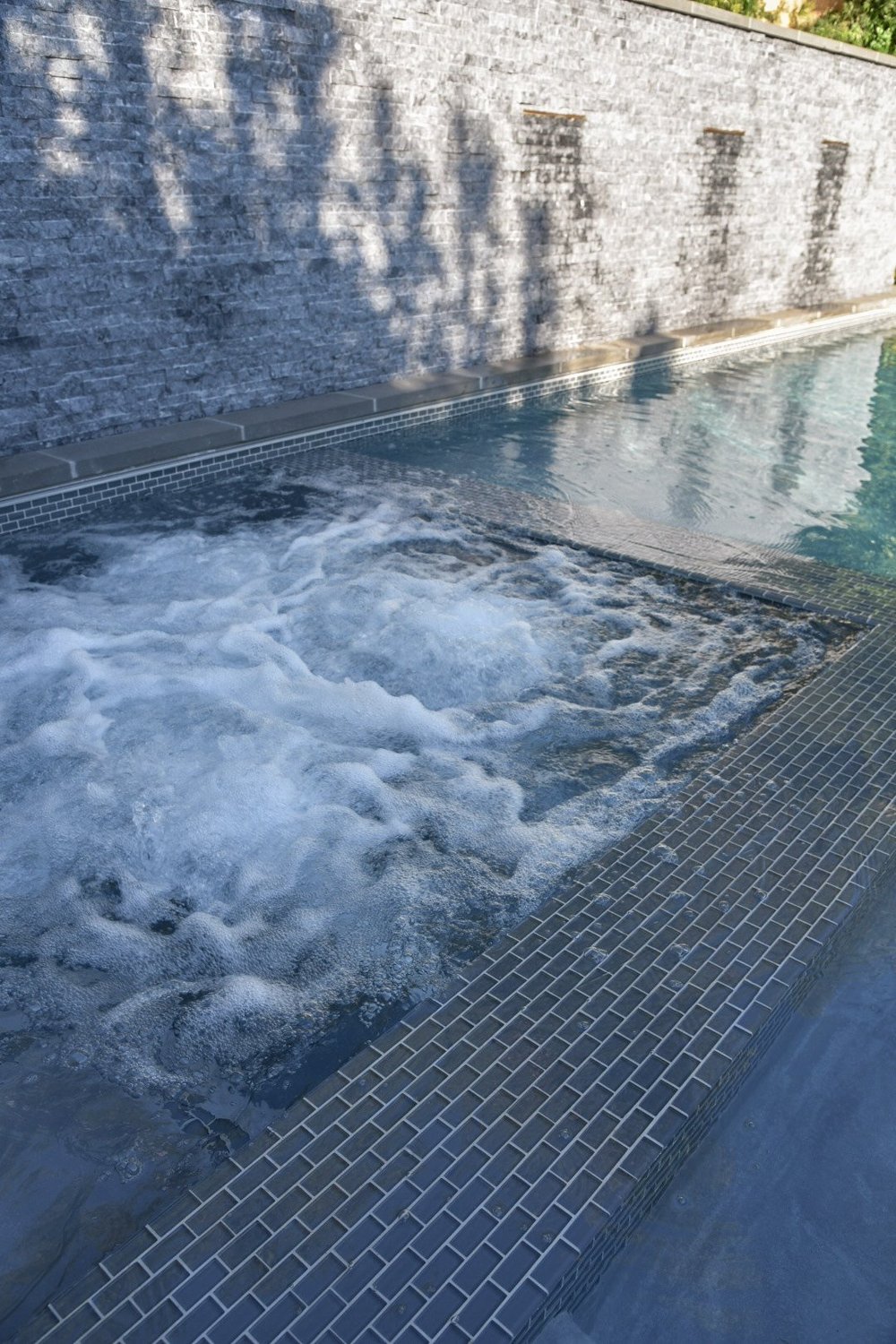 Bubbling spa water behind a tiled divider, with the adjoining pool and gray stone water wall beyond.