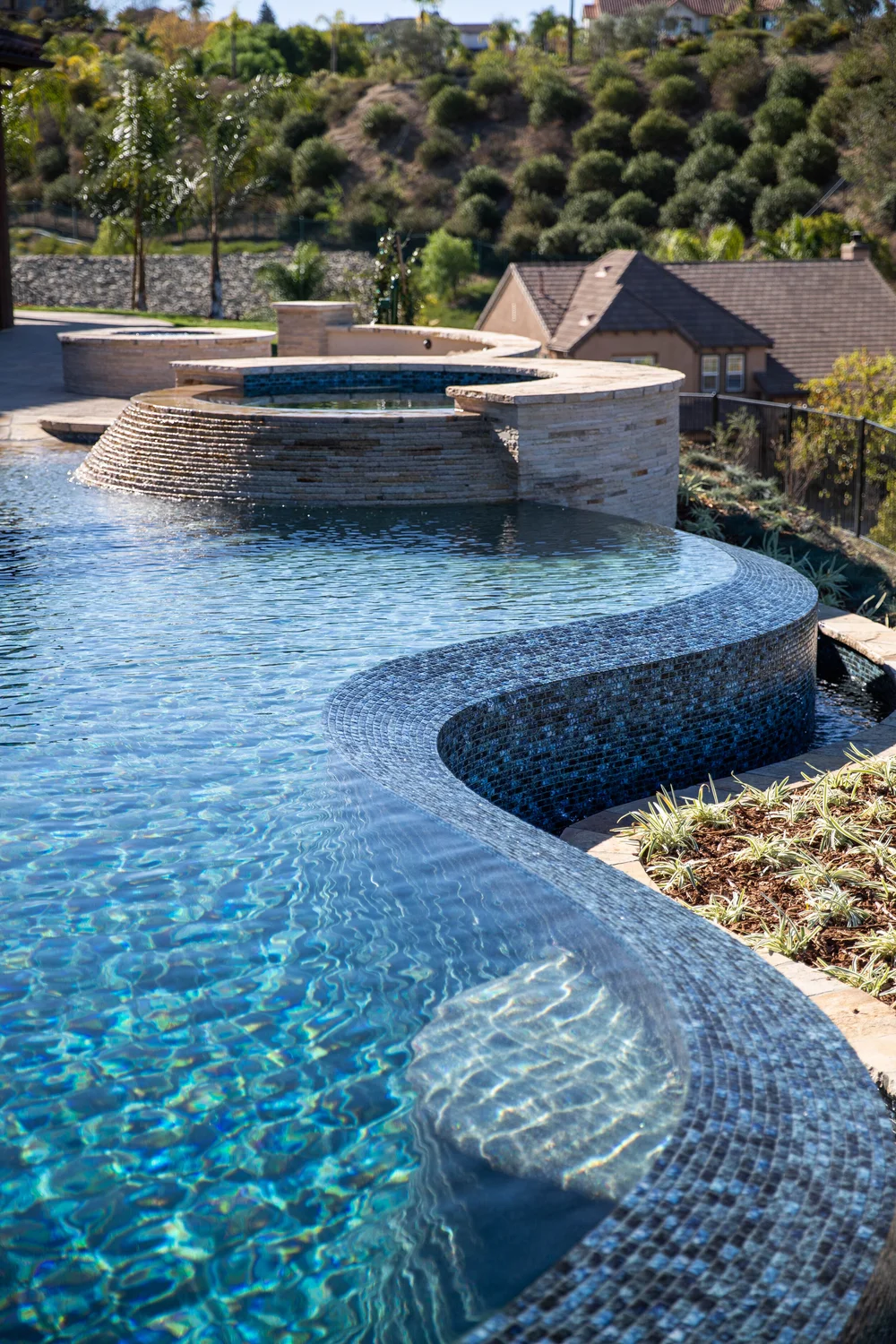 Blue mosaic infinity edge curving around a submerged stone, with a raised circular spa and hillside in the background.