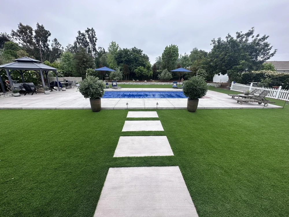 A straight stepping-stone path leads between two potted trees to a rectangular pool with a blue cover.