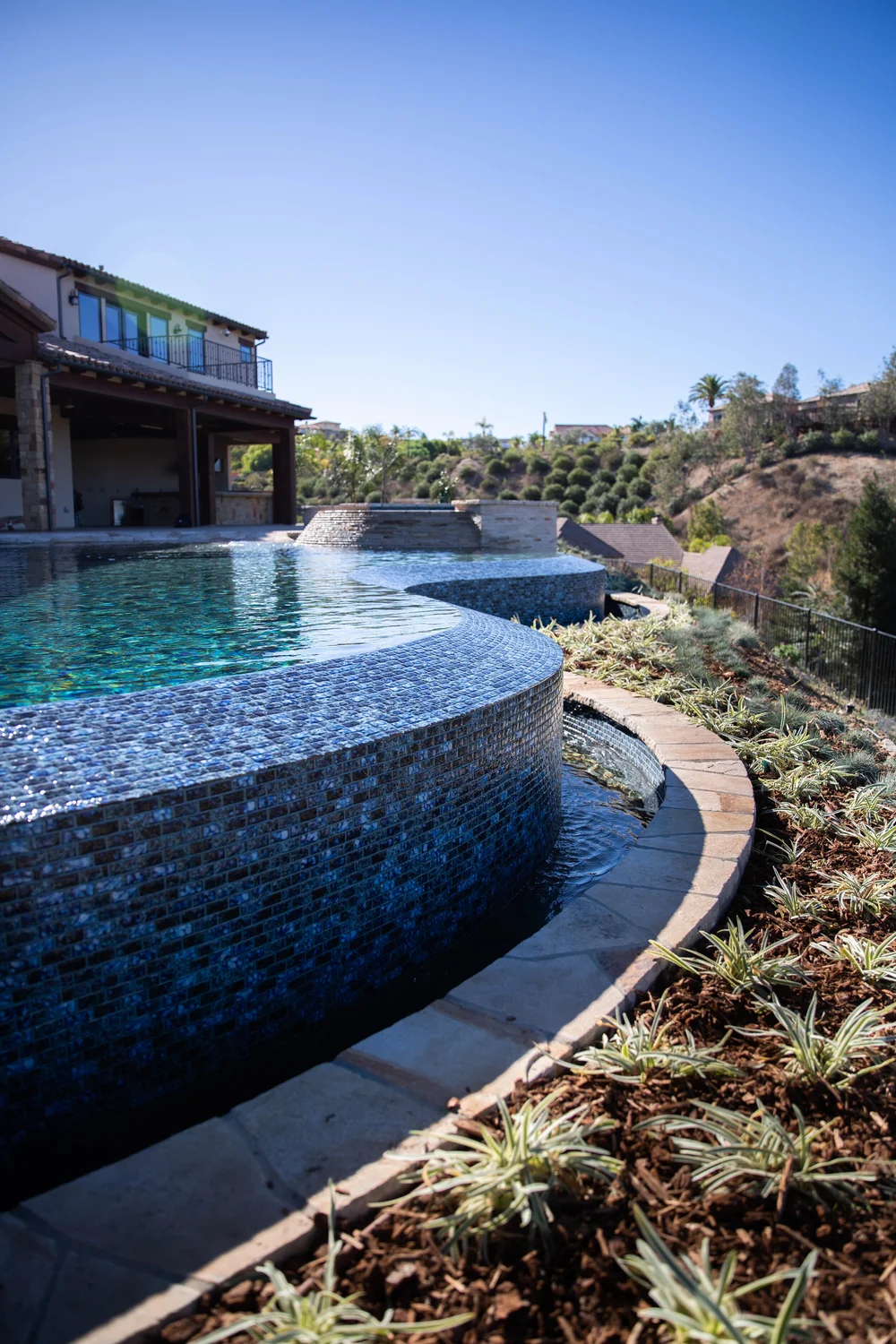 Curving blue mosaic infinity edge above a narrow catch basin, with a garden border and house beyond.
