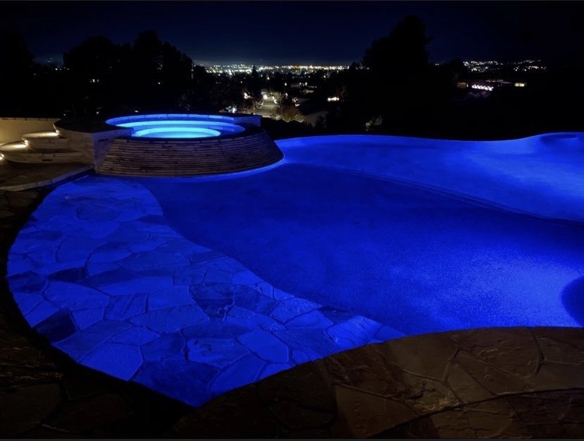 Freeform pool and raised circular spa glowing blue at night, with a shallow stone-lined entry in the foreground.