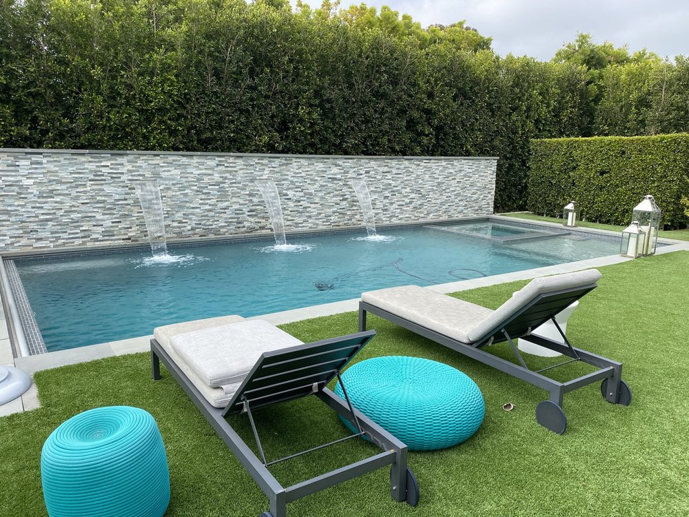 Two lounge chairs face a rectangular pool with arching water streams from a long textured wall.