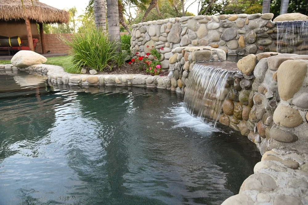 Two spillways cascade from a rounded-stone wall into a freeform pool beside flowers and a thatched seating area.