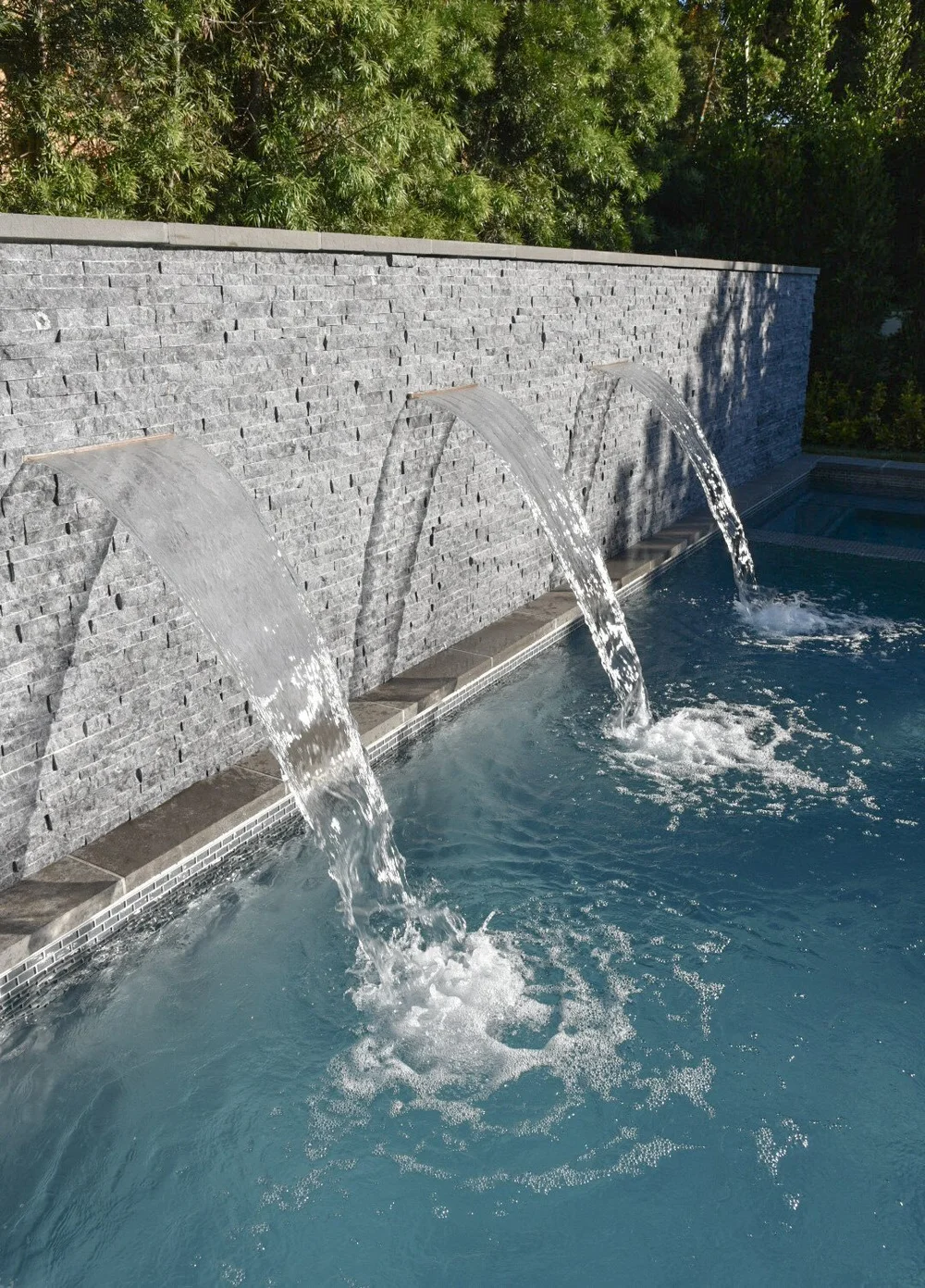 Angled close view of three spillways sending clear sheets of water from a gray stone wall into the pool.