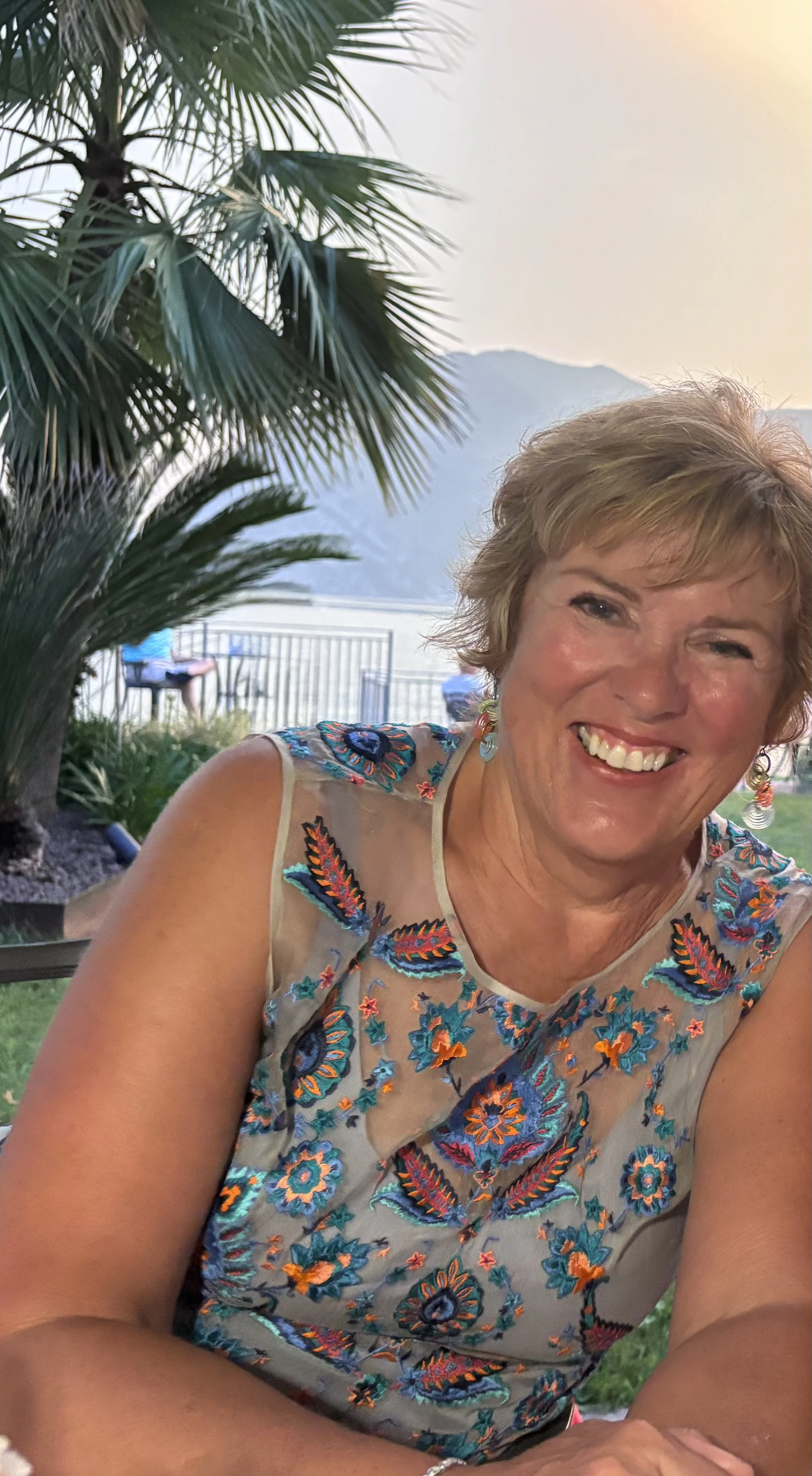 A woman smiling and sitting outdoors near a large palm tree with a lake, mountains, and chairs in the background.