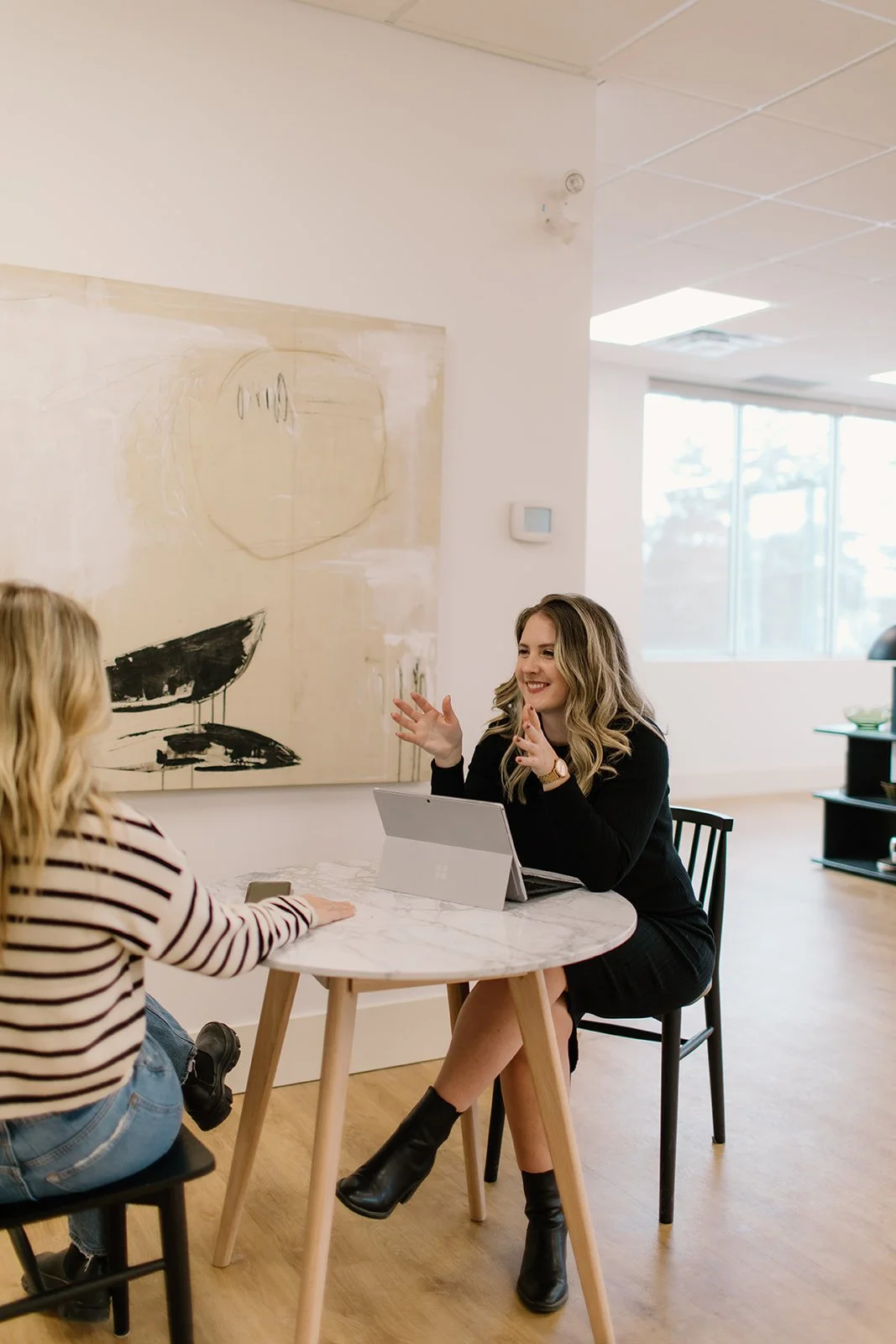 Two women sitting at a round table engaged in conversation in a modern office space, with a woman in a black outfit smiling and gesturing, a woman in a striped shirt holding a phone, and abstract artwork on the wall behind them.