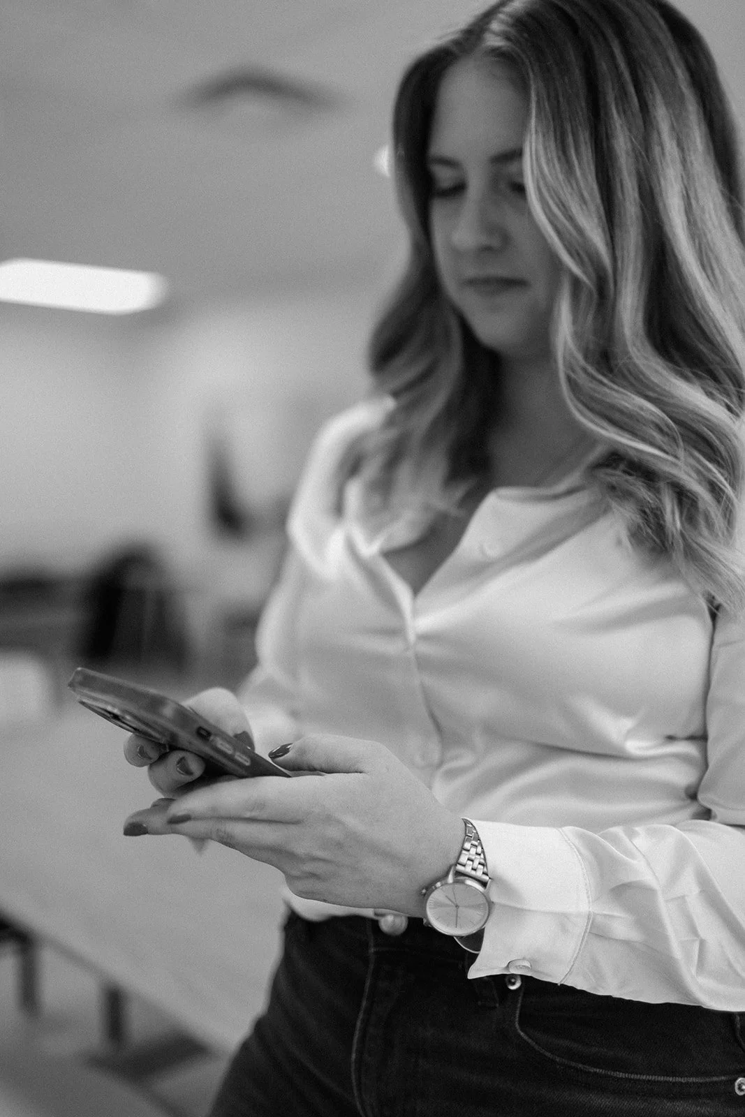 A woman with wavy hair wearing a white blouse and a watch on her left wrist, looking at her smartphone.