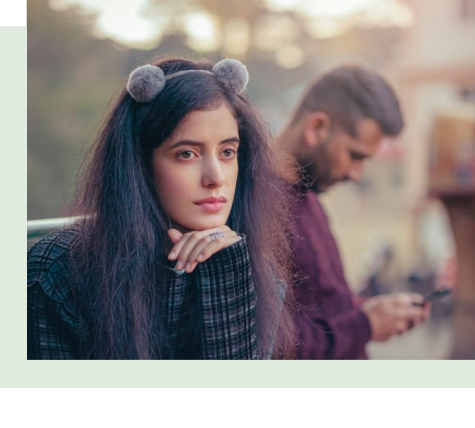 Woman looking away while her partner sits behind her on his phone, reflecting emotional distance and disconnection in a relationship.