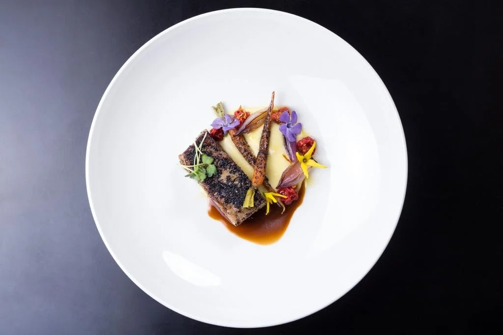Plato de comida gourmet con un filete con costra de pimienta negra, acompañado de vegetales decorativos y salsa, en un plato blanco