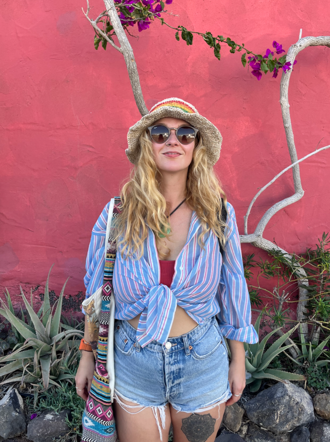 A woman with long blonde curly hair wearing sunglasses, a colorful striped shirt tied at the waist, denim shorts, a woven hat with a rainbow stripe, and a backpack, standing in front of a pink textured wall with plants and a twisted tree.