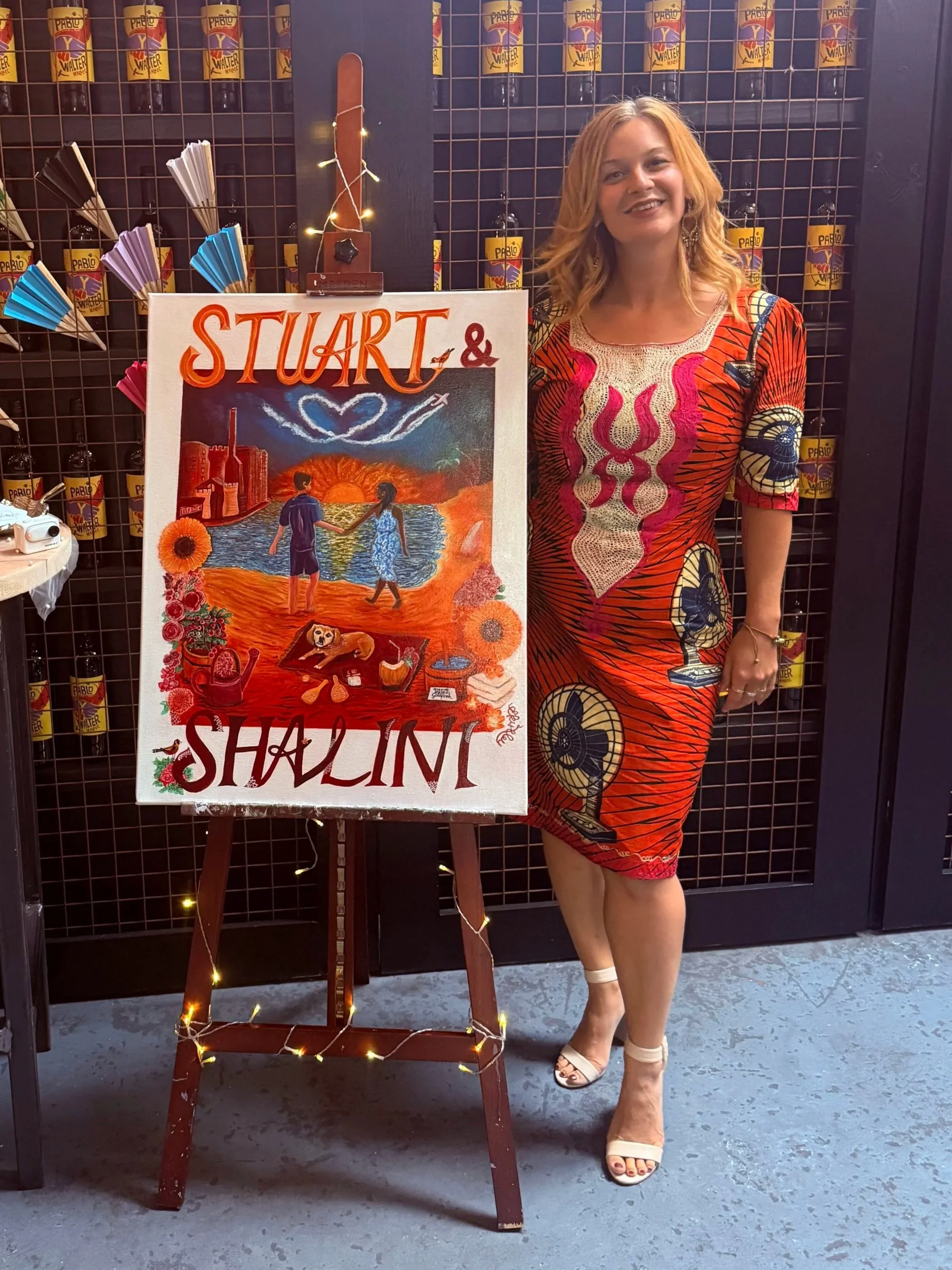 A woman in a colorful dress standing next to a painted welcome sign with a beach scene, holding a smile, surrounded by a dark grid backdrop and small bottles of Pablo Y Walter hot sauce on a shelf.
