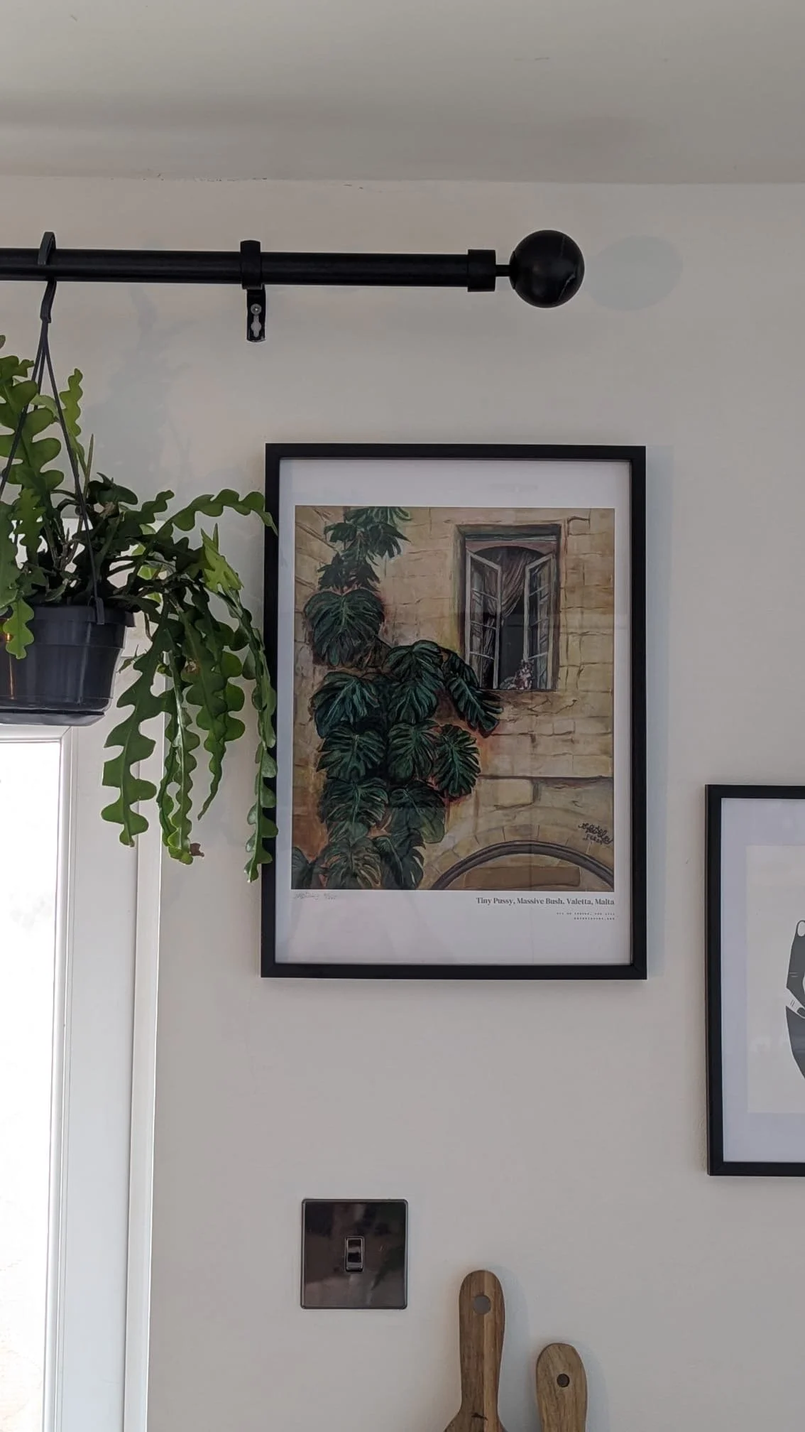 A framed artwork of a window with plants and a brick wall behind it hangs on a white wall, with a hanging plant to the left and kitchen utensils below.