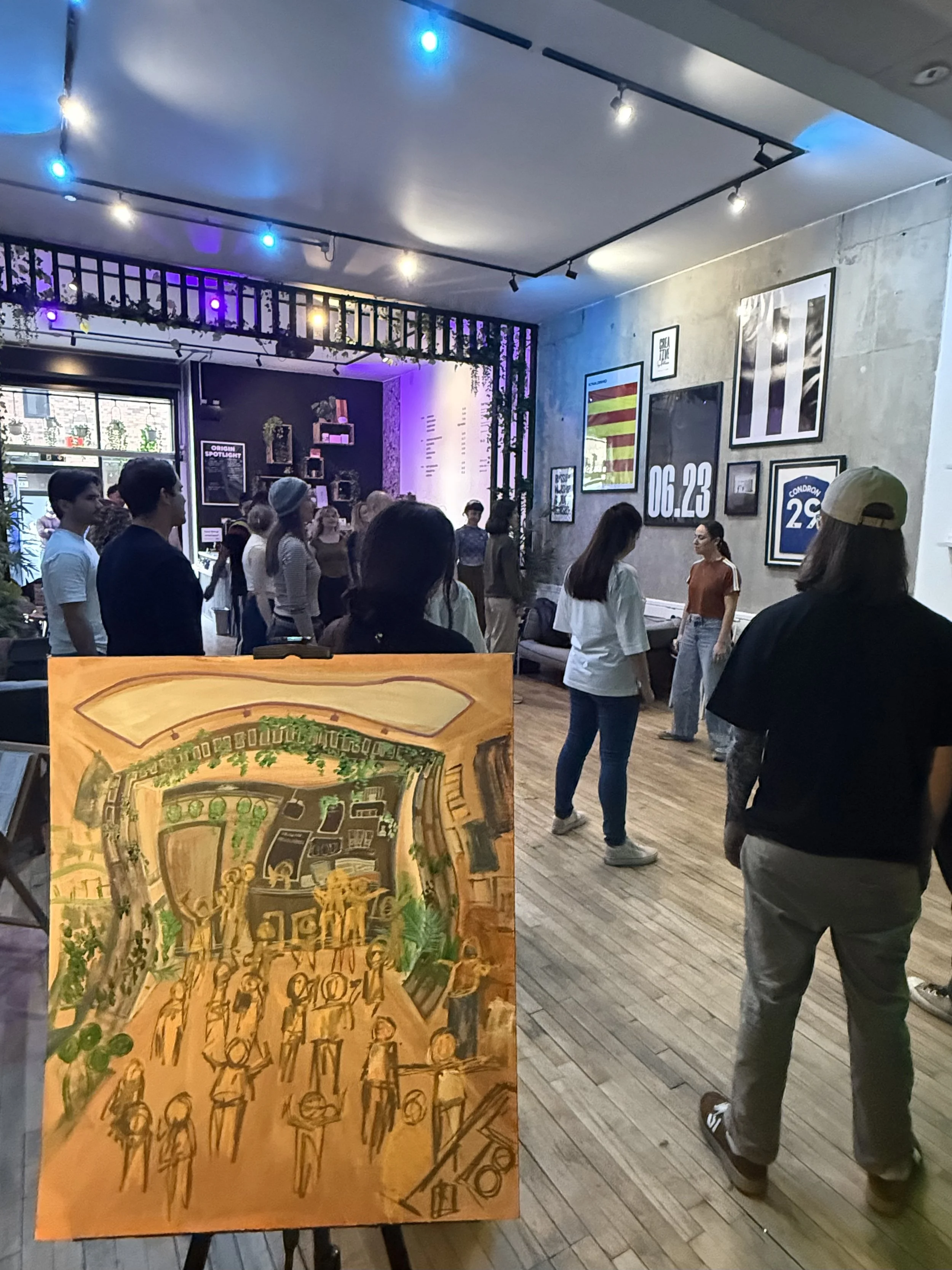 A crowd of people in an art gallery or museum with framed artwork on the wall, and a colorful painting on an easel in the foreground.