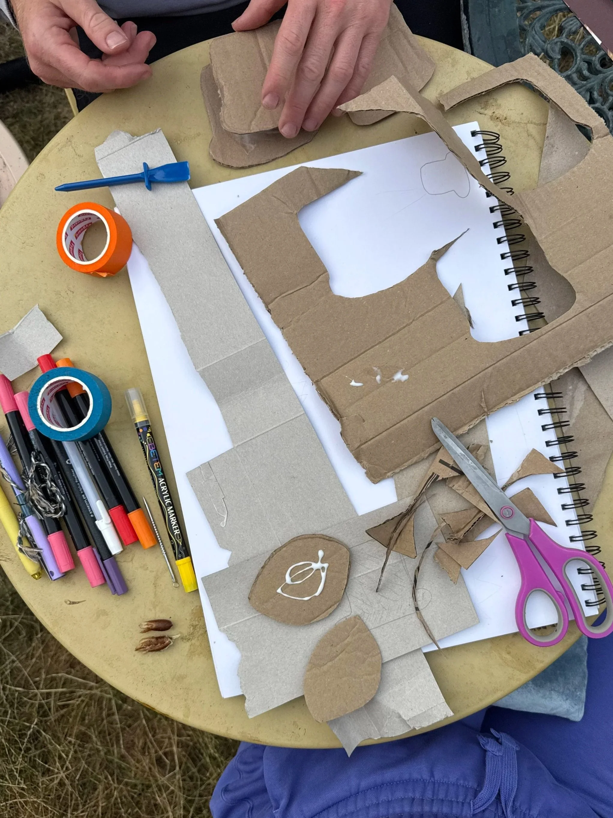 A person using cardboard and crafting tools to make a project, surrounded by markers, scissors, and tape on a yellow table, with a notebook underneath.