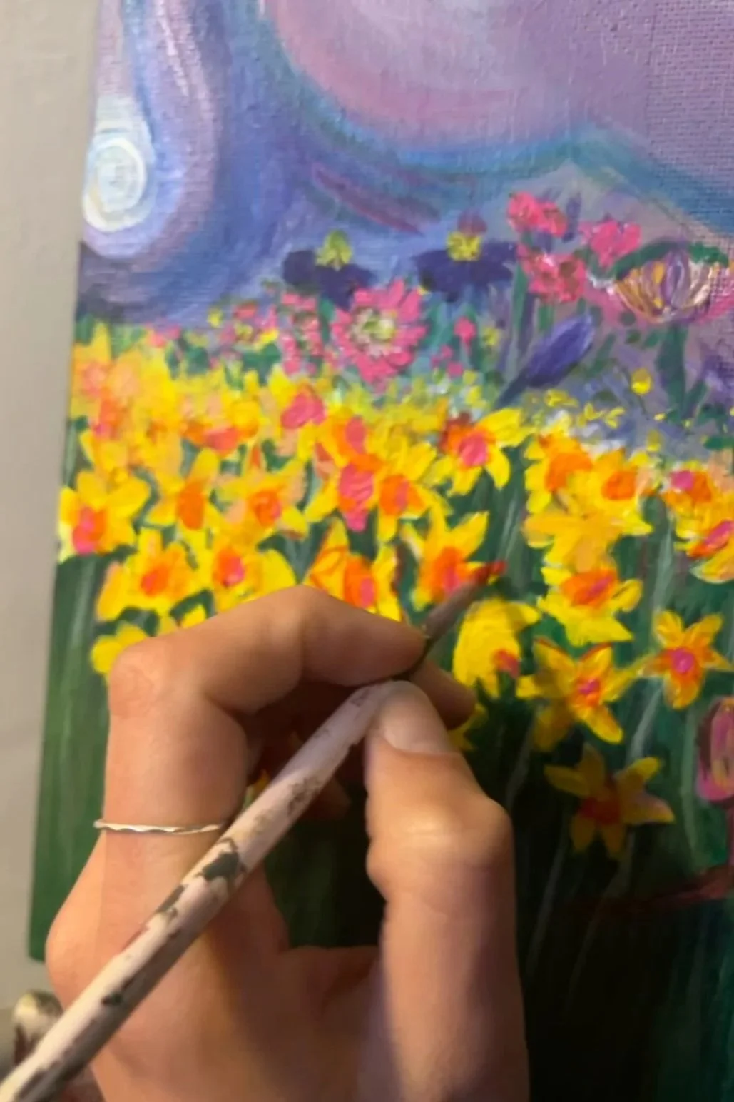 A person is painting a colorful floral scene with bright yellow, pink, purple, and blue flowers on a canvas. The person is holding a paintbrush and is in the process of creating the artwork.