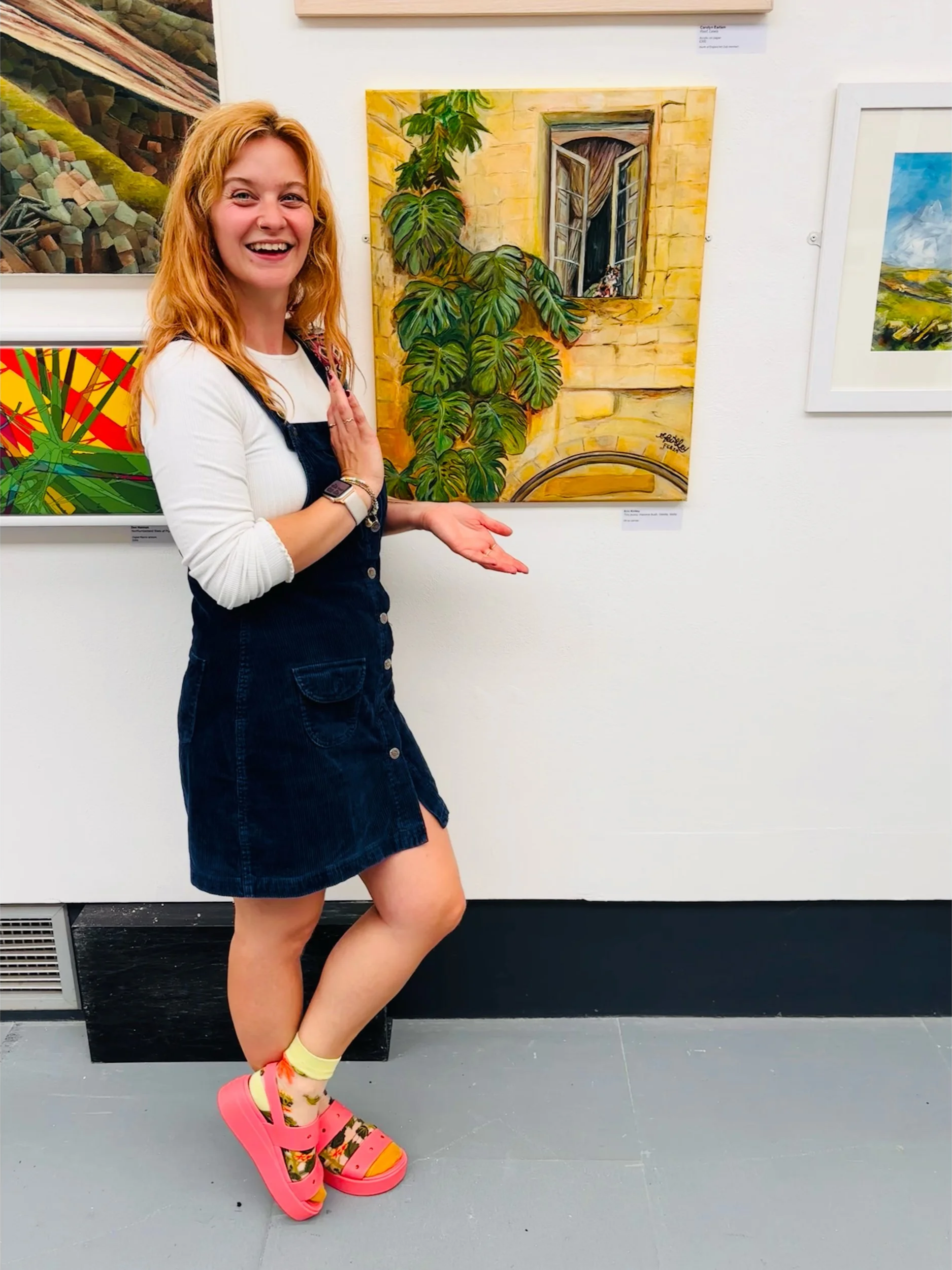 A woman with red hair, wearing a white long-sleeve top, black denim dress, and colorful platform sandals, standing in an art gallery, smiling and gesturing towards a colorful painting of a window with greenery.