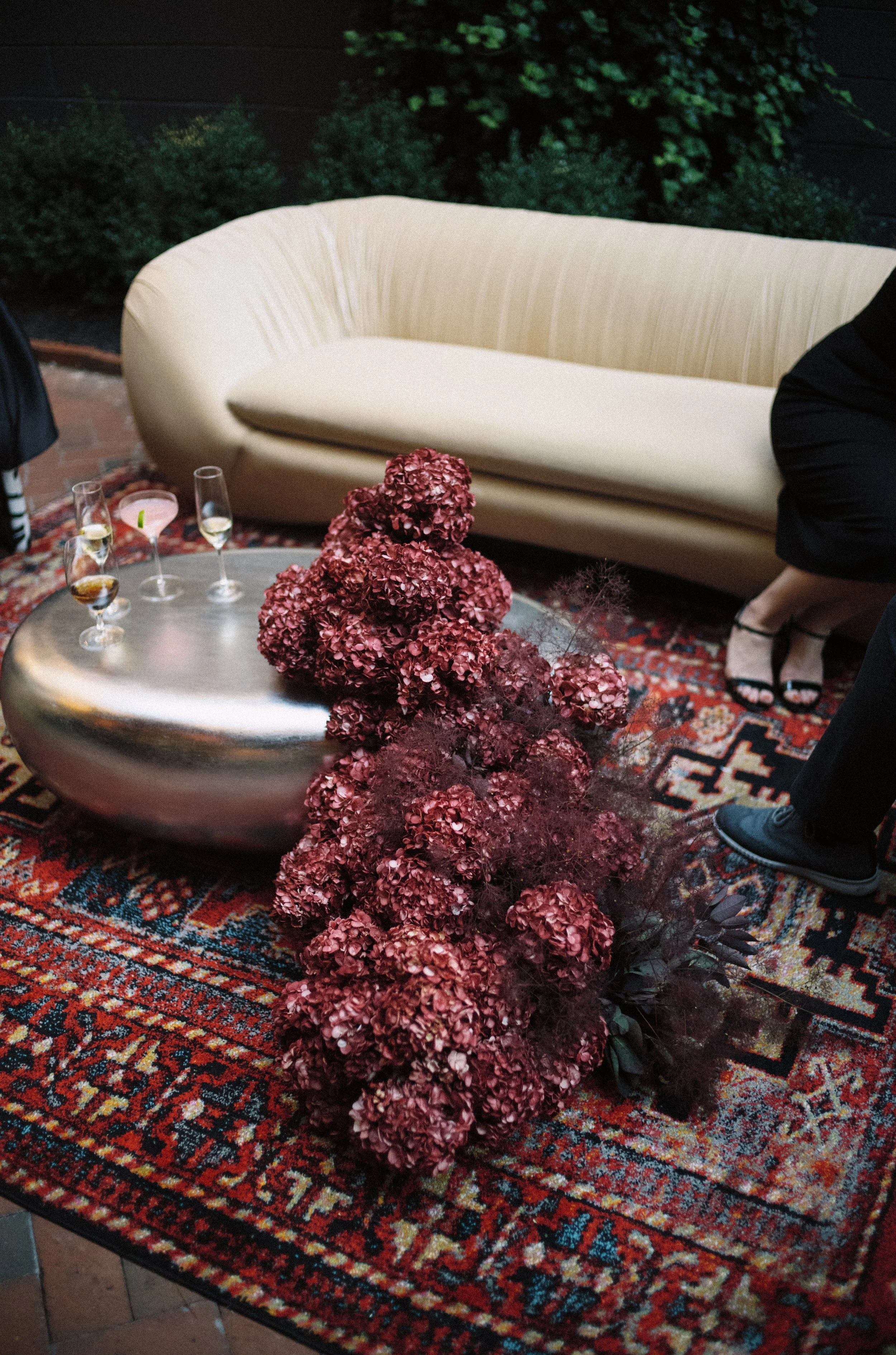 Floral arrangement with a mass of dark red hydrangeas, dark colored greenery, and decorative elements on a brightly colored rug, with a silver table and white couch in the background.