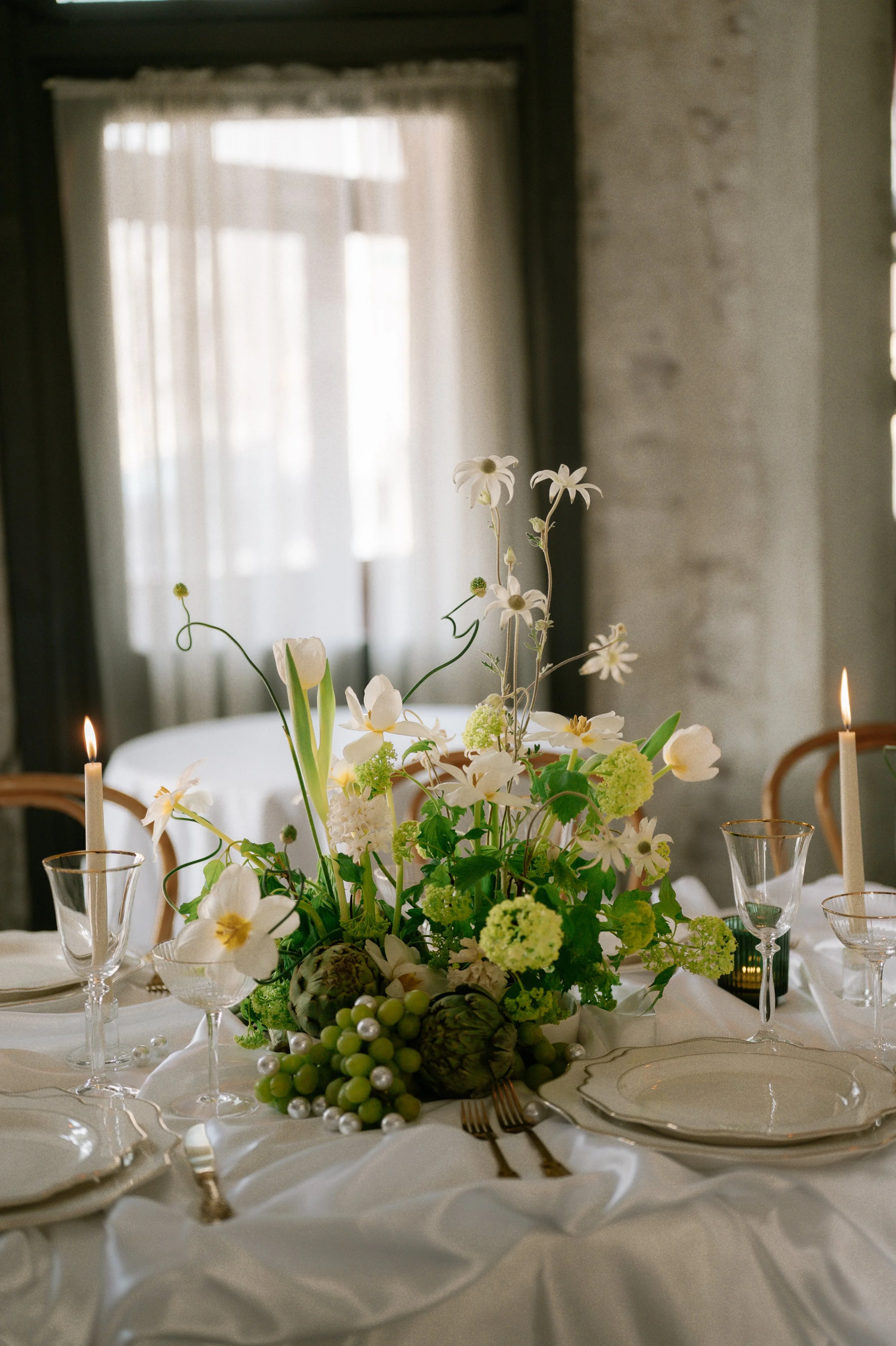 Elegant dinner table with a white floral centerpiece, candles, glassware, and plates, set in a bright room with curtains.