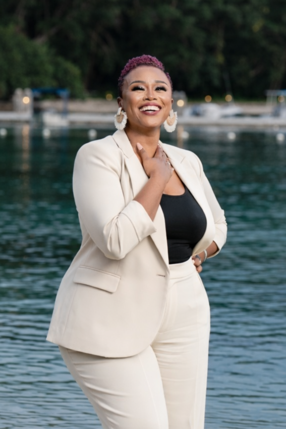 Smiling woman in a cream suit with black top and large white earrings by a body of water, with trees in the background.