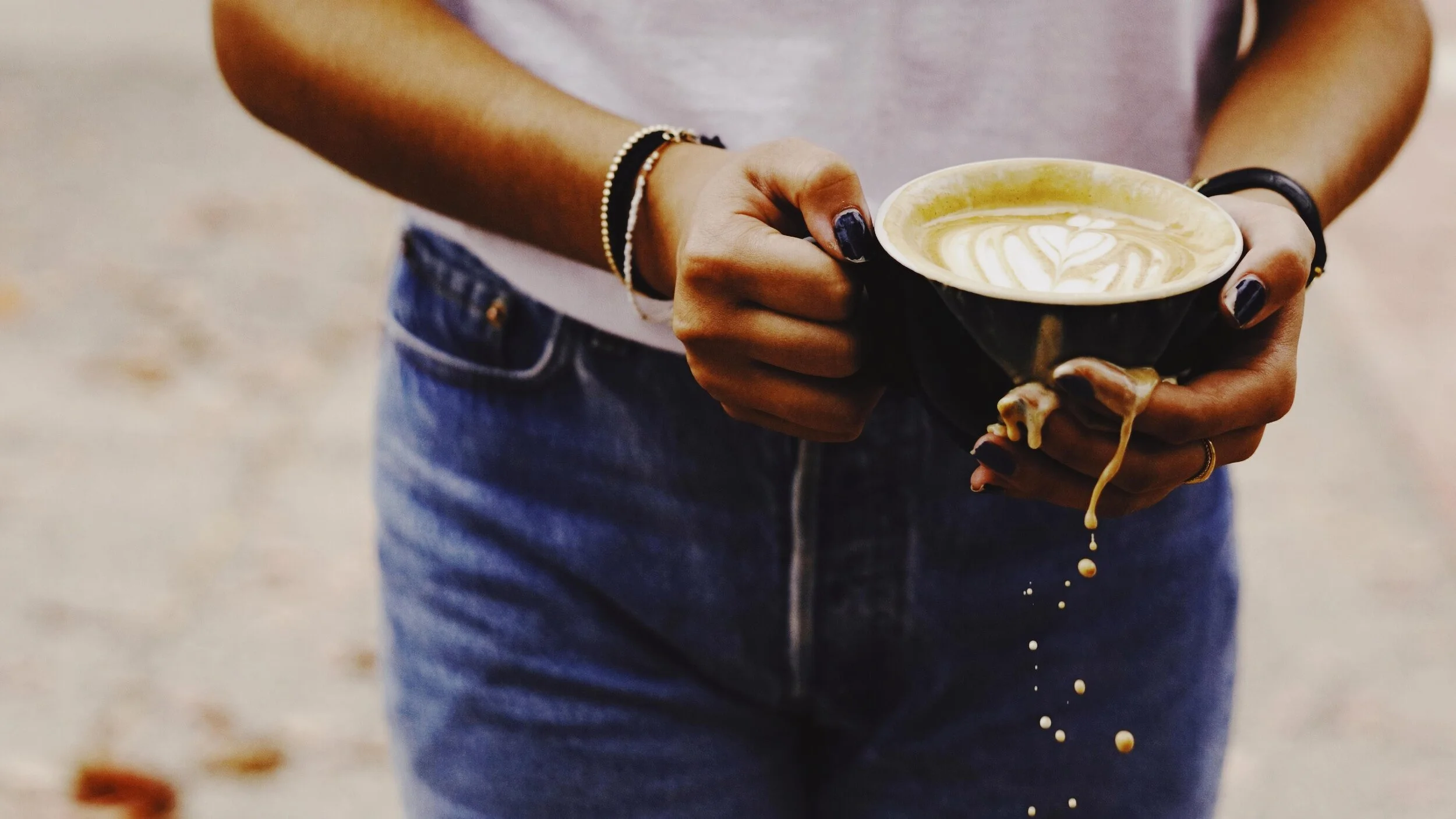 Person holding a black cup of matcha latte with latte art, wearing rings and bracelets, dressed in a gray top and blue jeans, with spilled matcha on hands.