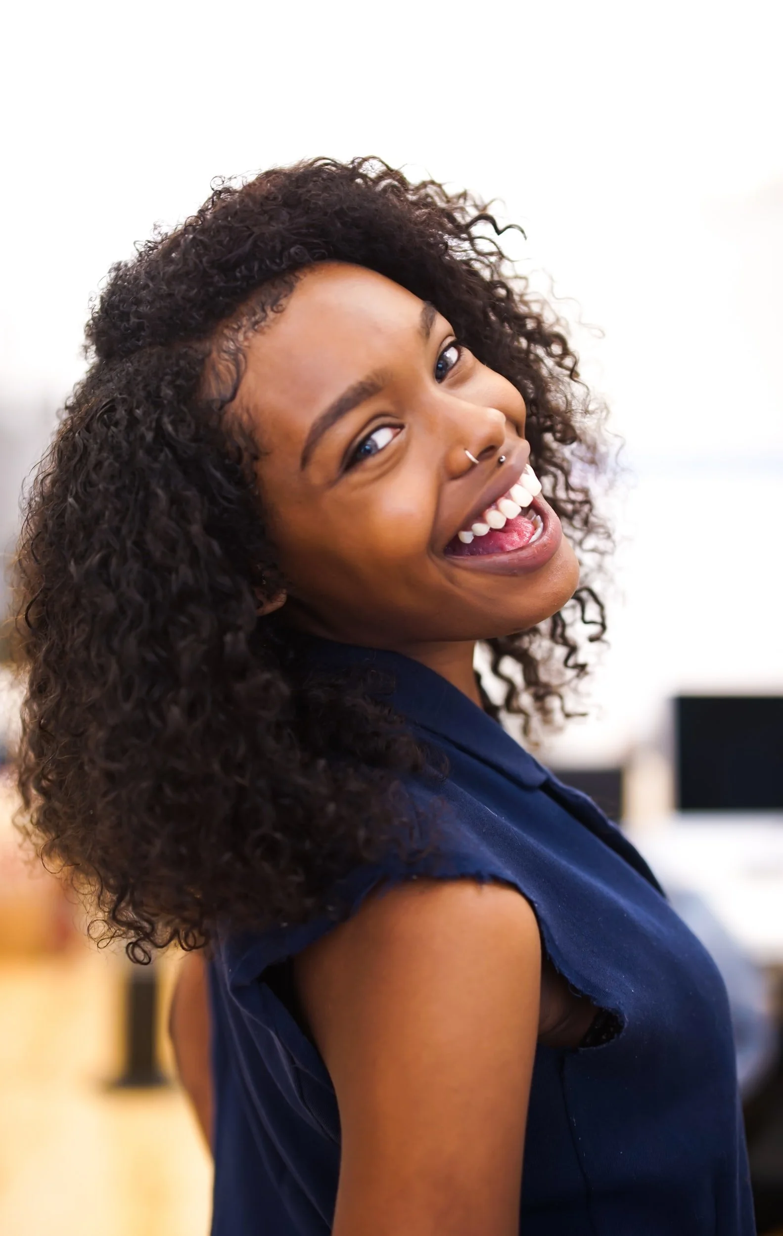 A young woman with curly hair smiling and looking over her shoulder, wearing a navy blue sleeveless top, in an indoor setting.