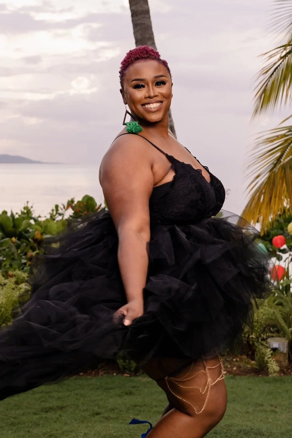 A woman with short pink curly hair, wearing a black lace dress with a tulle skirt, stands outdoors near a palm tree with a scenic ocean view in the background. She is smiling and holding her dress.