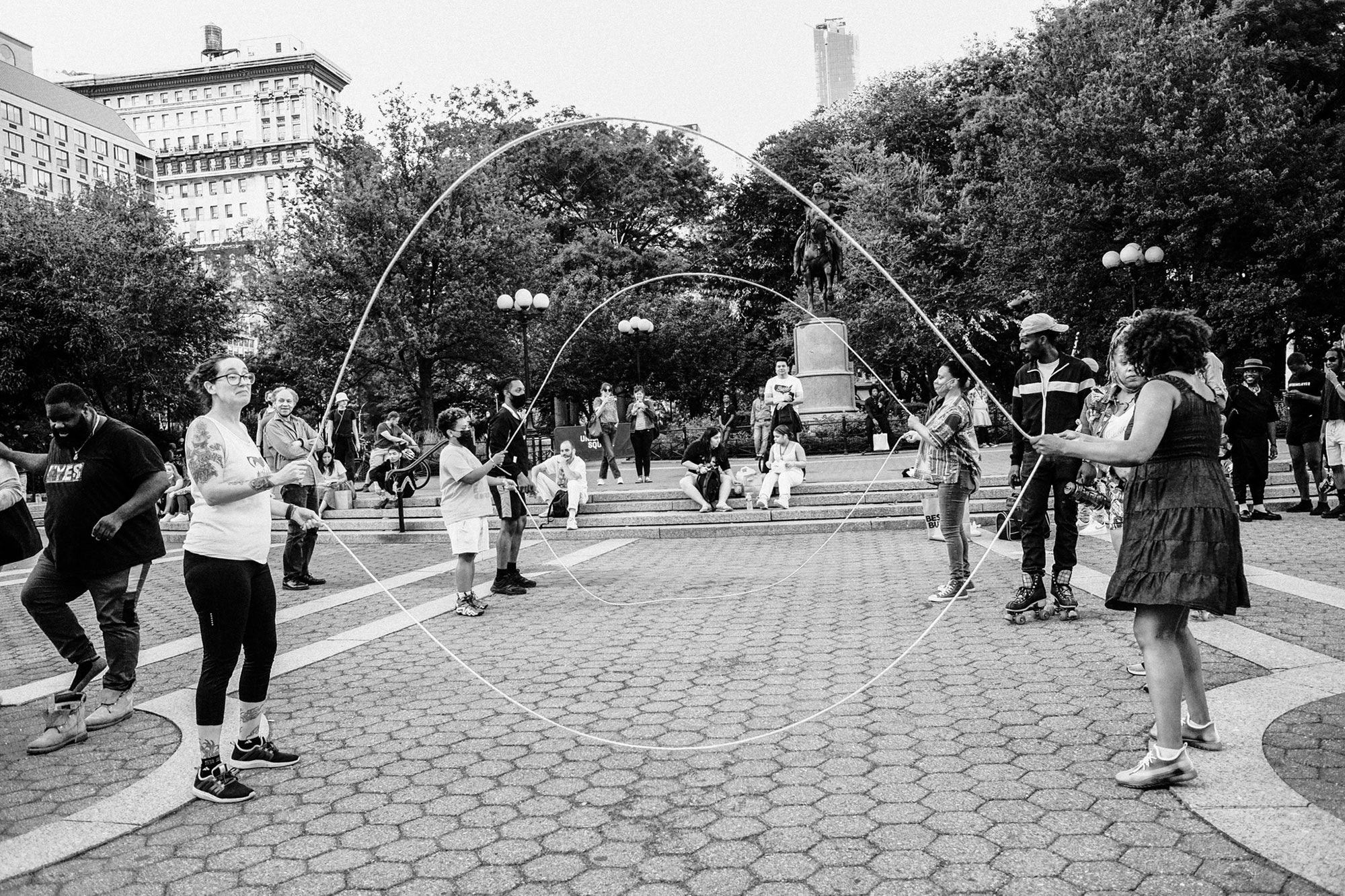 Black and white photo of many people gathered around a city square as multiple people jump double Dutch.