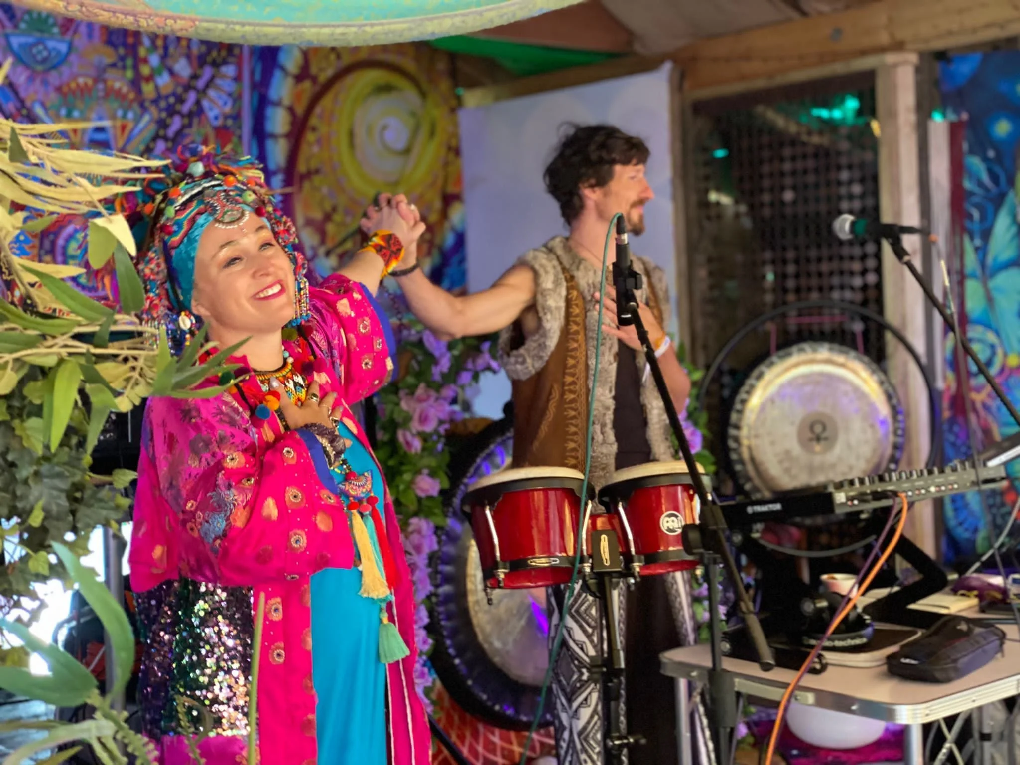 Two performers on stage, one woman in colorful traditional attire with jewelry and a headwrap, smiling and holding hands with a man in tribal-inspired clothing, standing near musical instruments including drums and a gong, with vibrant, decorative background.