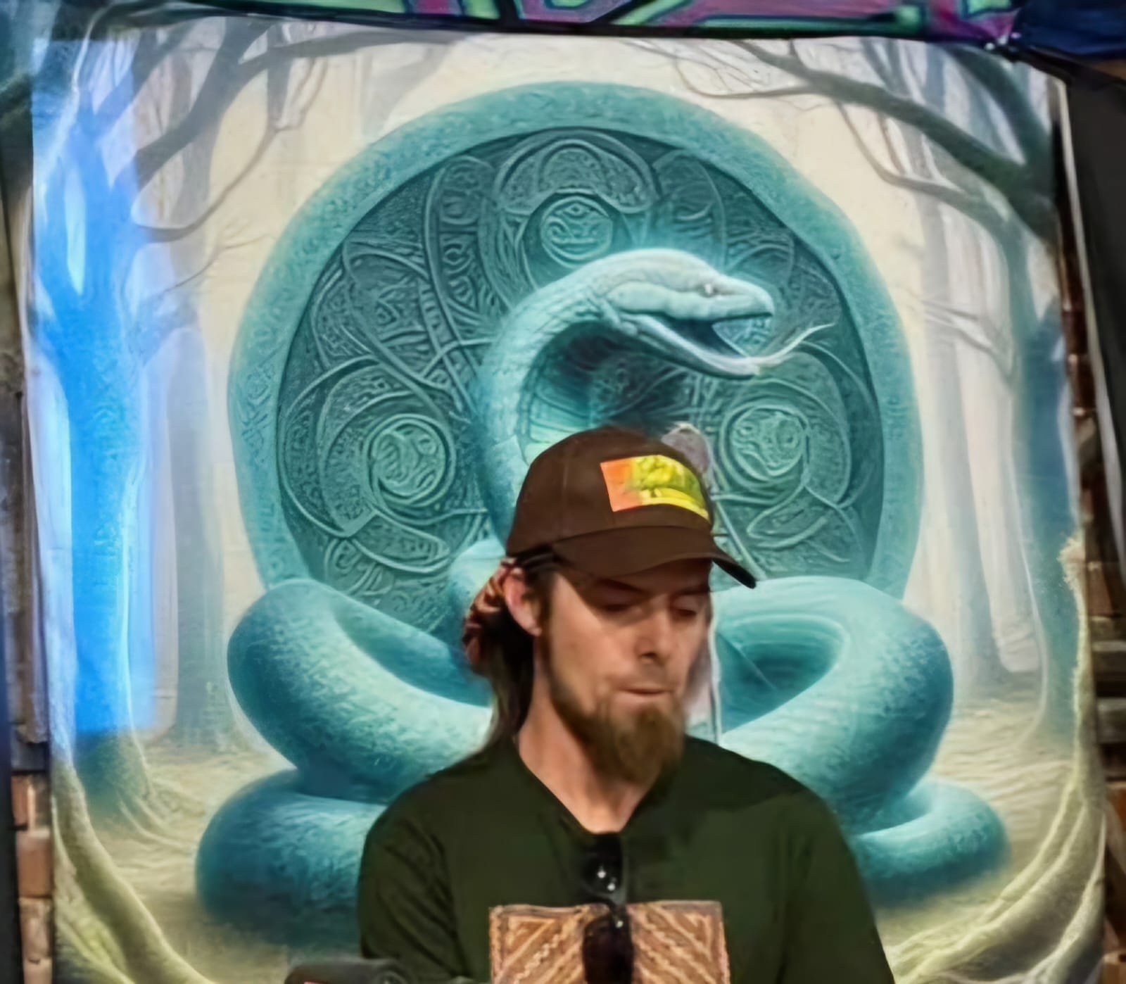 A man with a beard wearing a brown cap and black shirt stands in front of a fantasy-style tapestry featuring a large snake coiled around a circular symbol with intricate designs.
