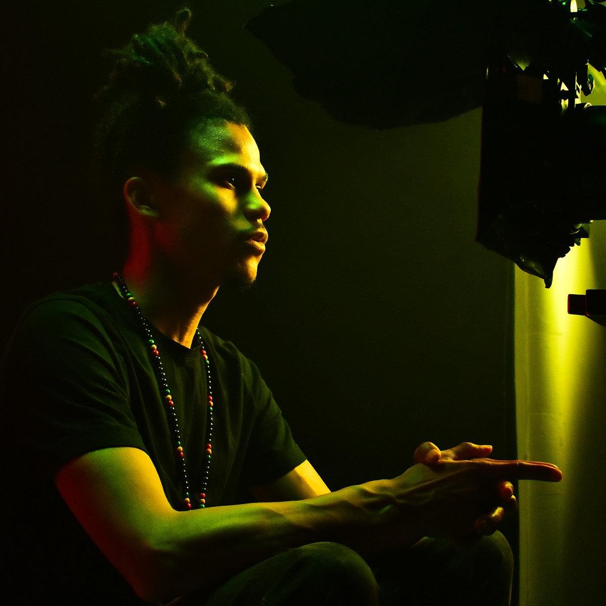 A young person with dreadlocks, wearing a black shirt and beaded necklace, is illuminated by greenish lighting, sitting in a dark room with a camera or filming equipment nearby.
