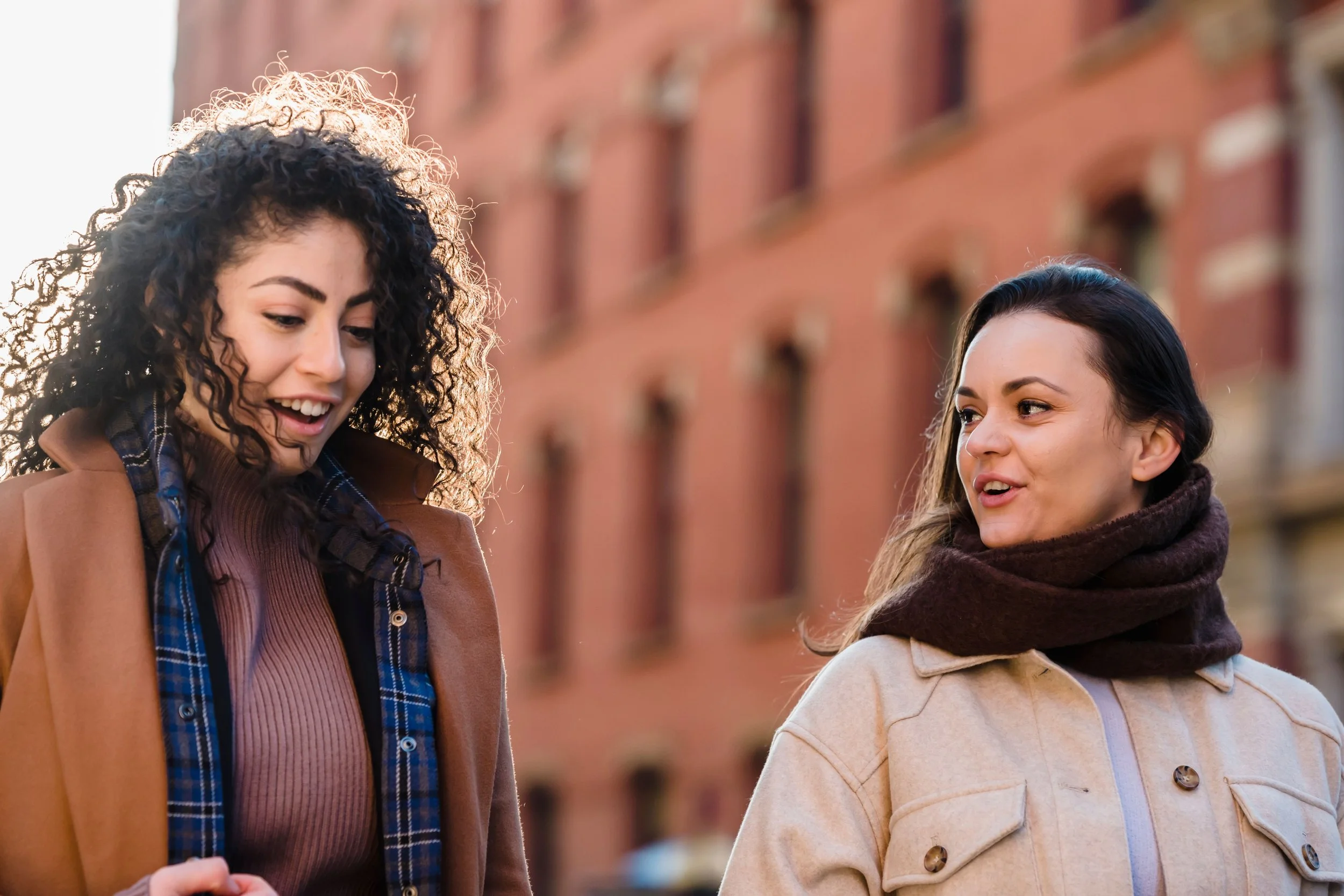 Two women standing outdoors in front of a red brick building, conversing and smiling.