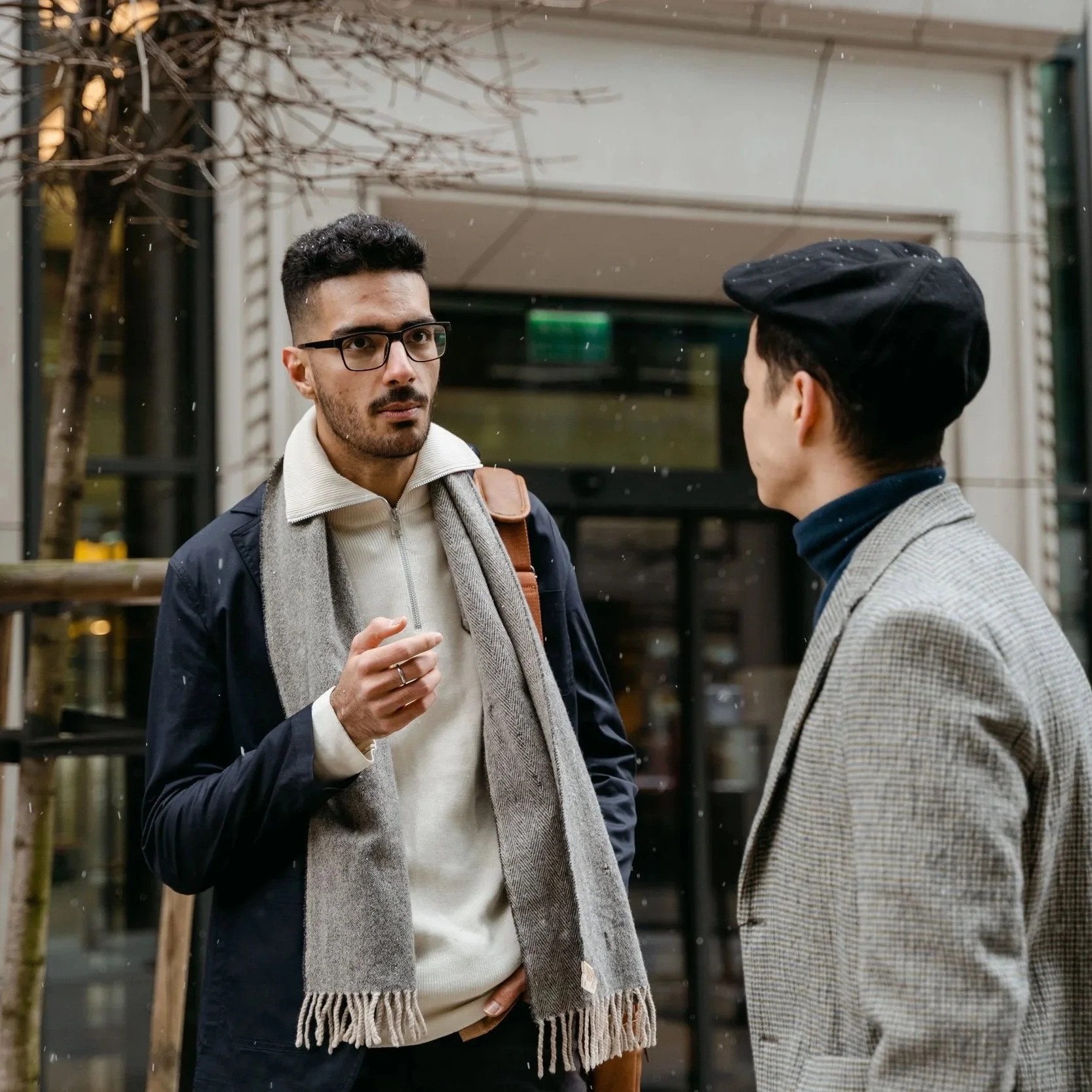 Two men having a conversation outside near a building with glass doors and a leafless tree, one wearing glasses and a dark jacket, the other wearing a gray blazer and a black beret.