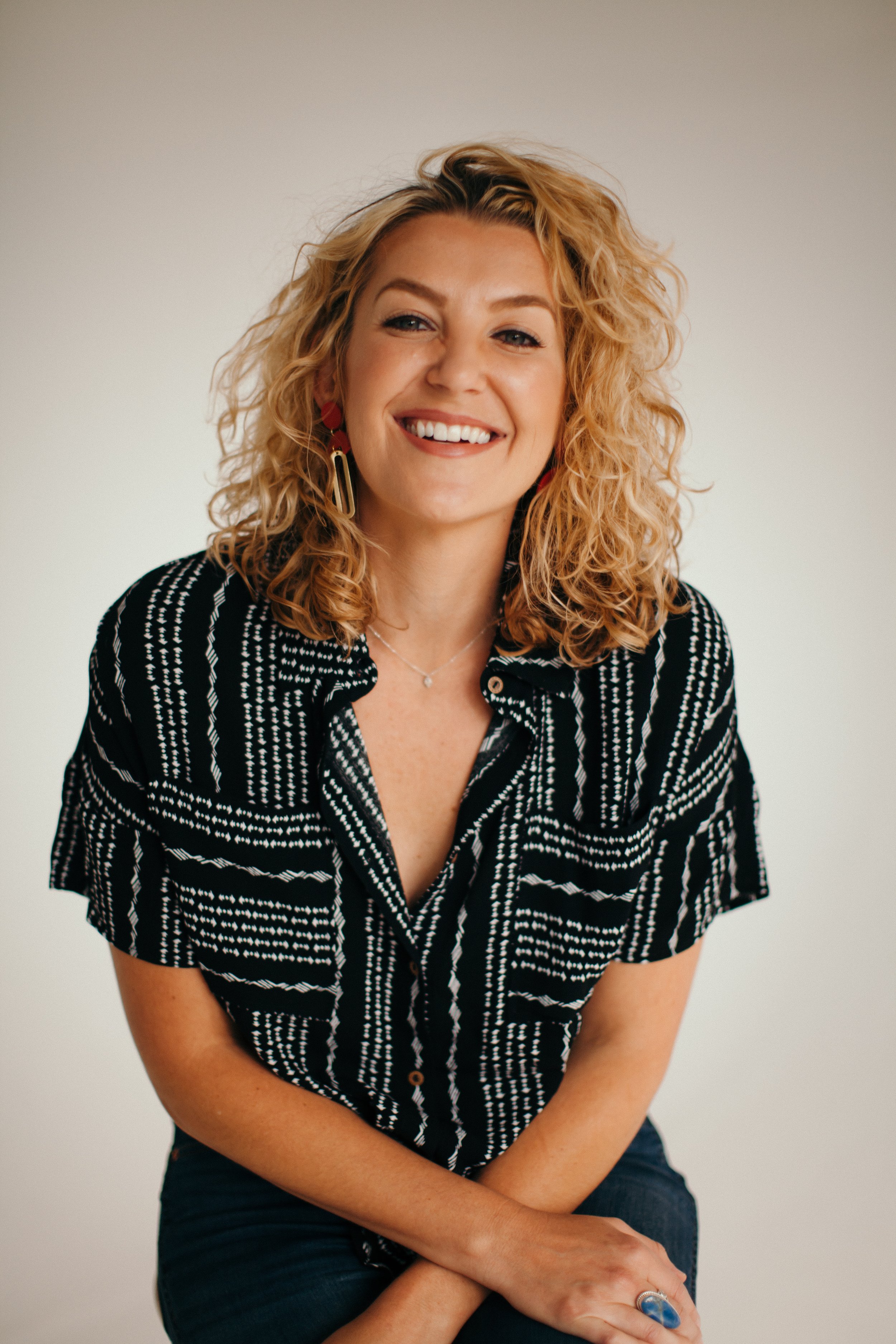 Portrait of a smiling woman with curly blonde hair, wearing a black and white patterned short-sleeve shirt, sitting against a plain light-colored background.