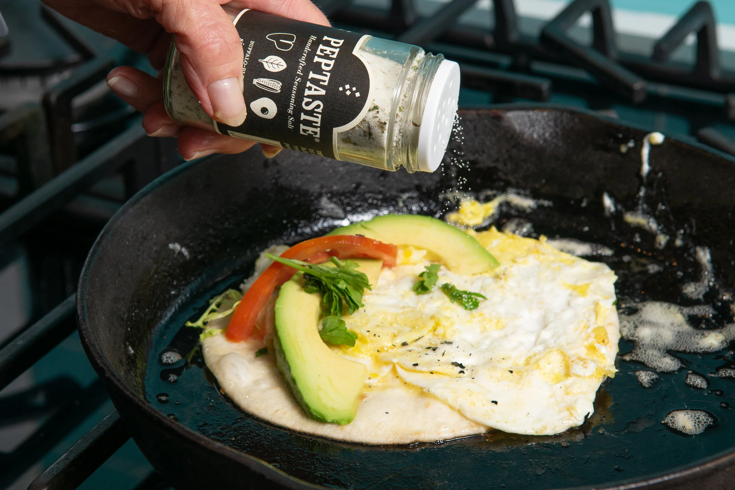 Open Peptaste® bottle sprinkled by hand over eggs with red pepper, avocado, and herbs in a cast iron skillet.